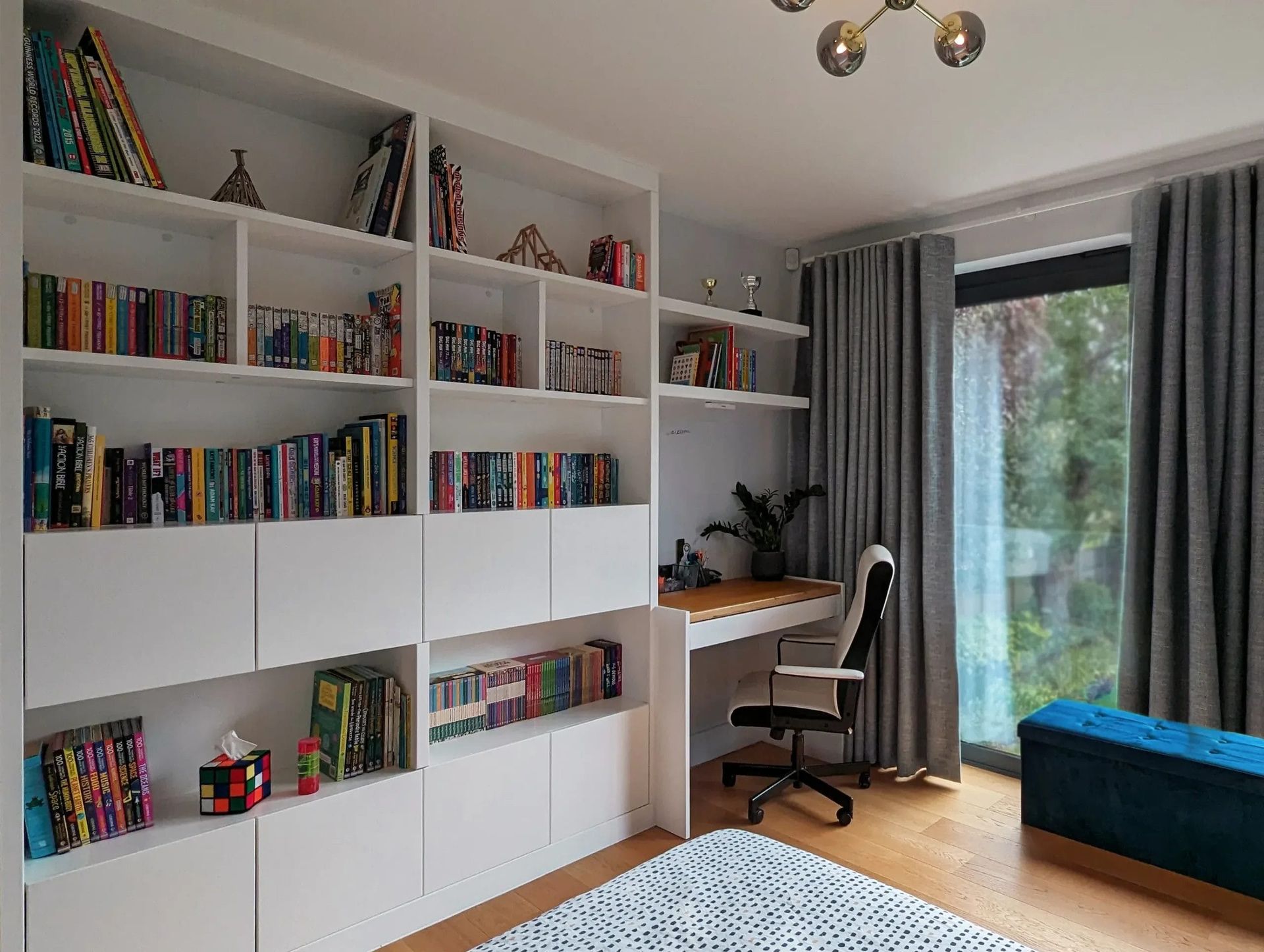 White bookshelves flank a desk and window in a room. Books, decorative items, and curtains are visible.