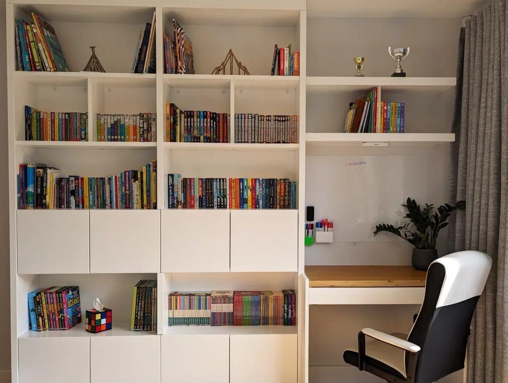 White bookshelf with books and desk. A plant, chair, and curtain are also visible.