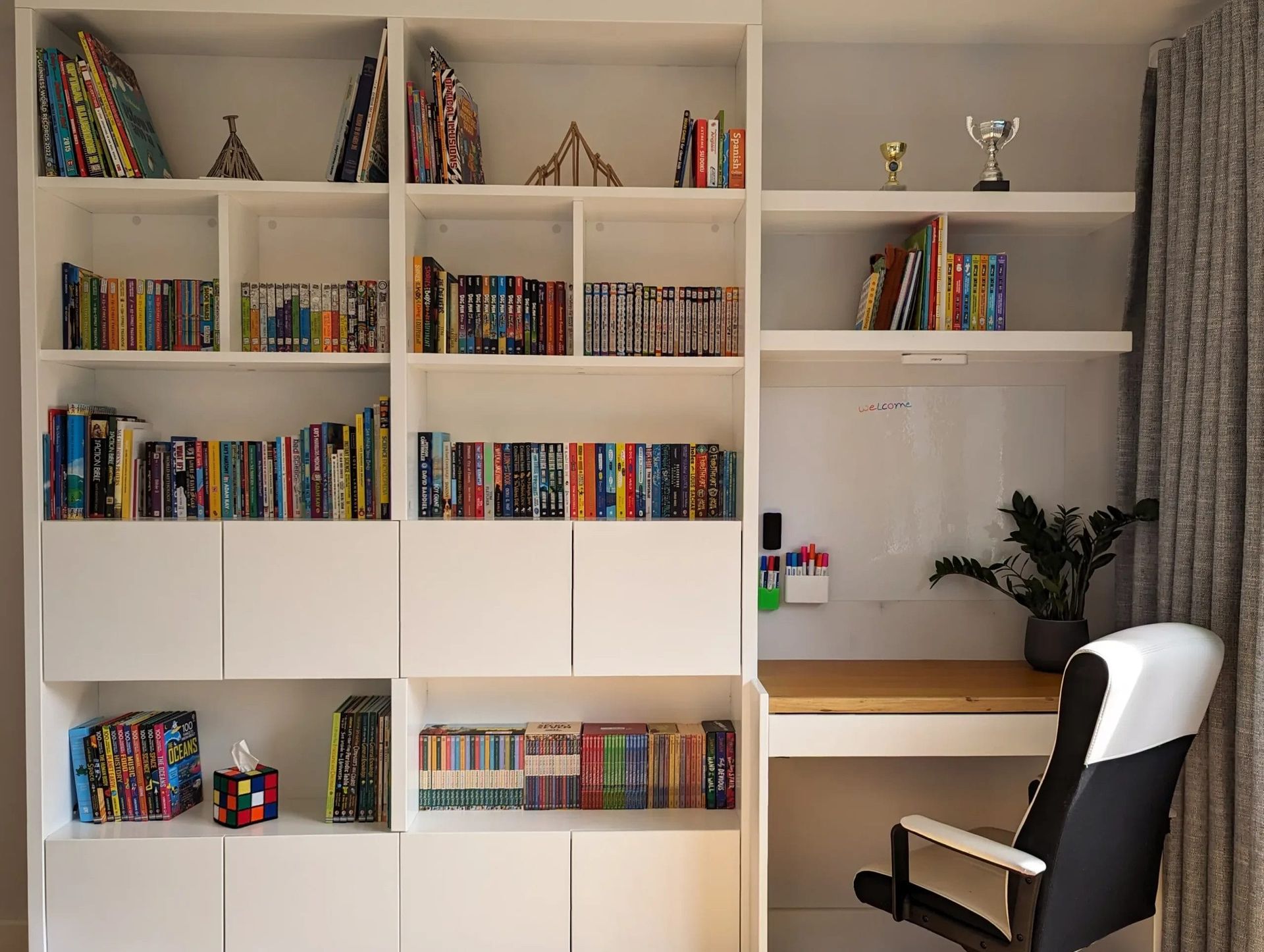 White built-in bookcase with books, DVDs, and a desk, next to a curtain.