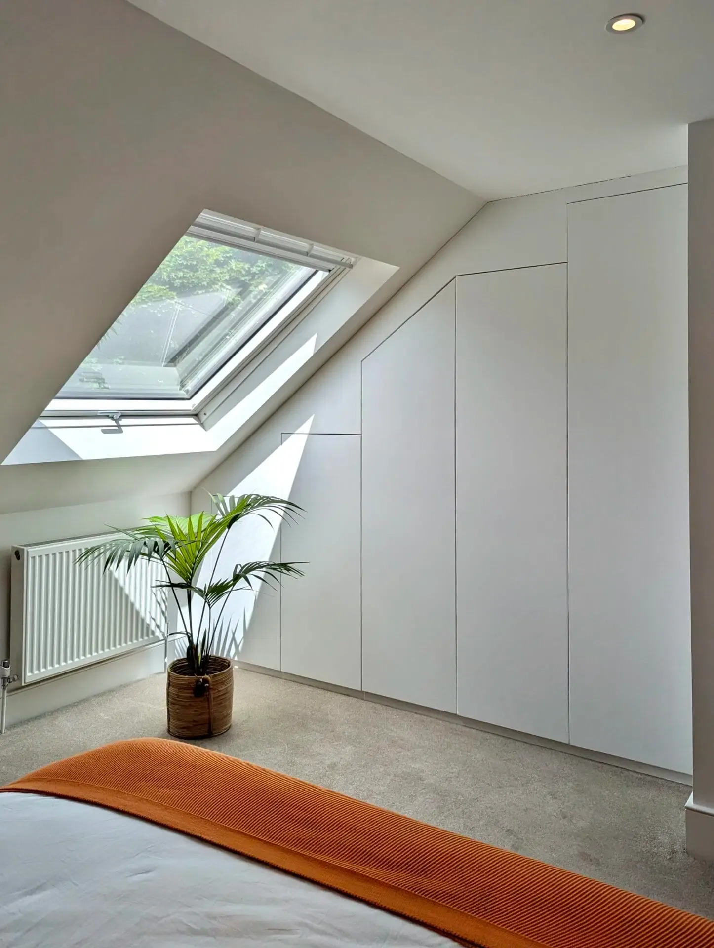 Bedroom with angled ceiling, skylight, white wardrobe, plant, radiator, and orange blanket.