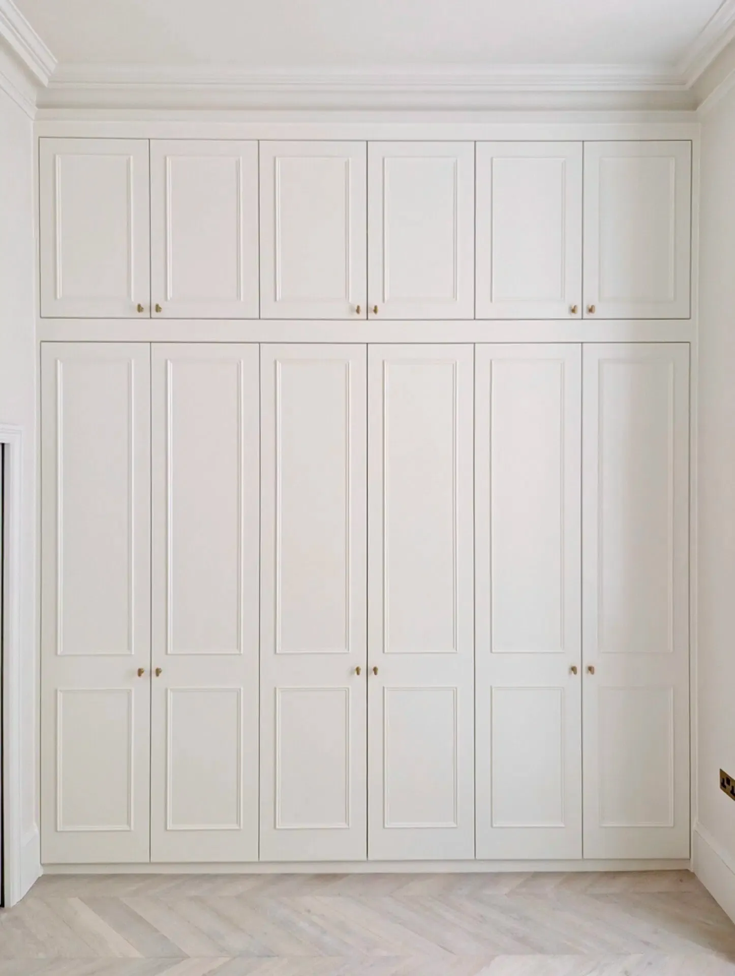 White built-in wardrobe with multiple doors and upper cabinets, against a white wall and light wood floor.