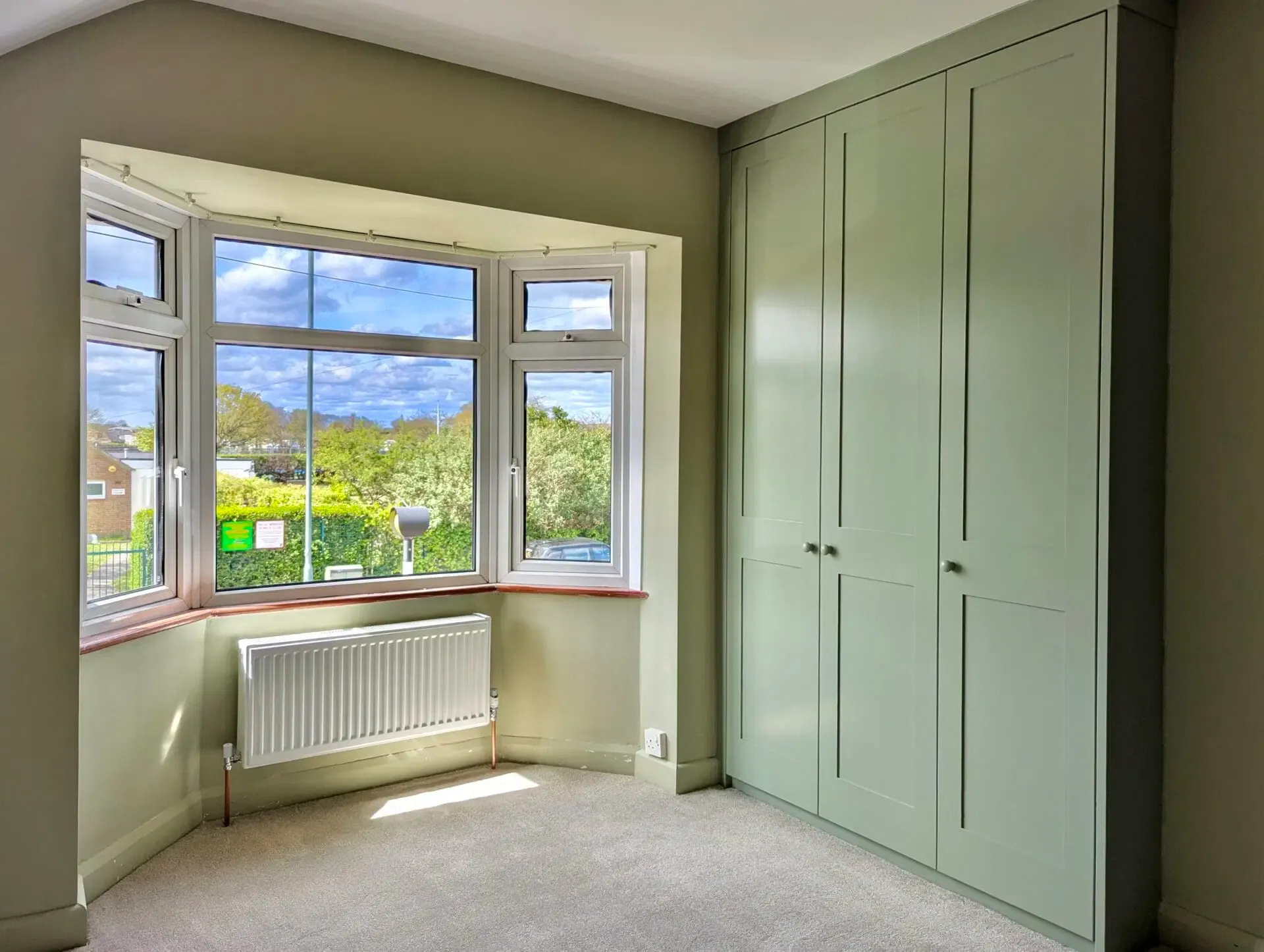 Bedroom with bay window and sage green built-in closet. Sunny, with view of trees and sky.