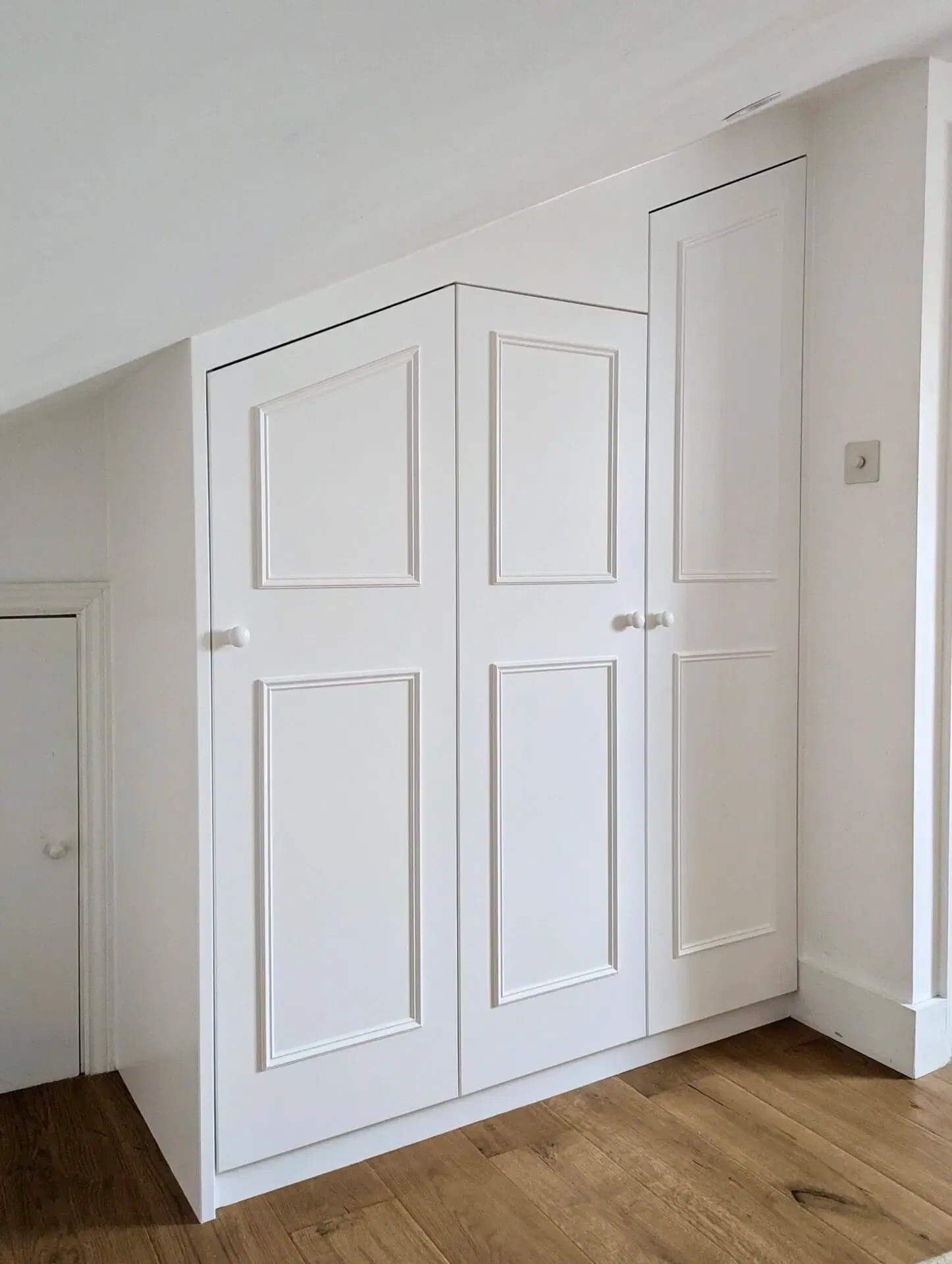 White built-in wardrobe with angled top and panelled doors against white walls and wood floor.