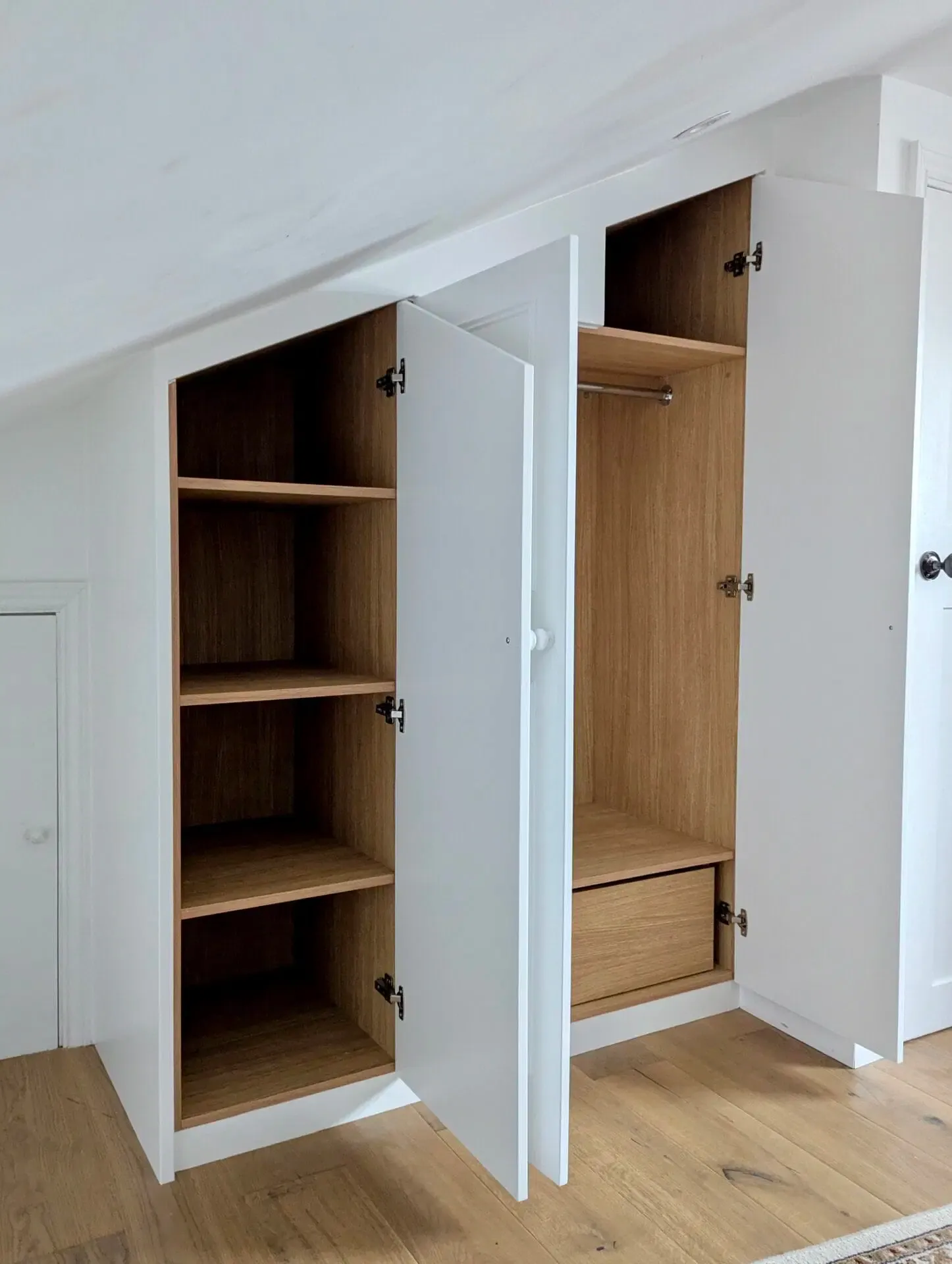 White closet with open doors, shelves and a hanging rod, built-in to angled wall.