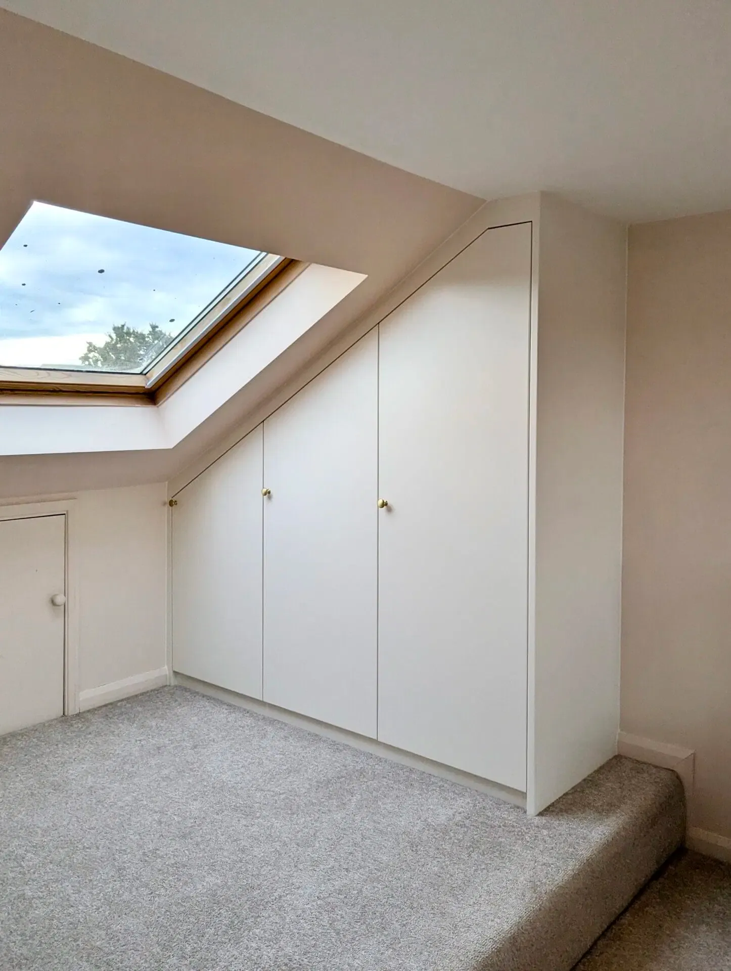 White built-in storage cabinets under a sloped ceiling next to a skylight in a room with gray carpet.
