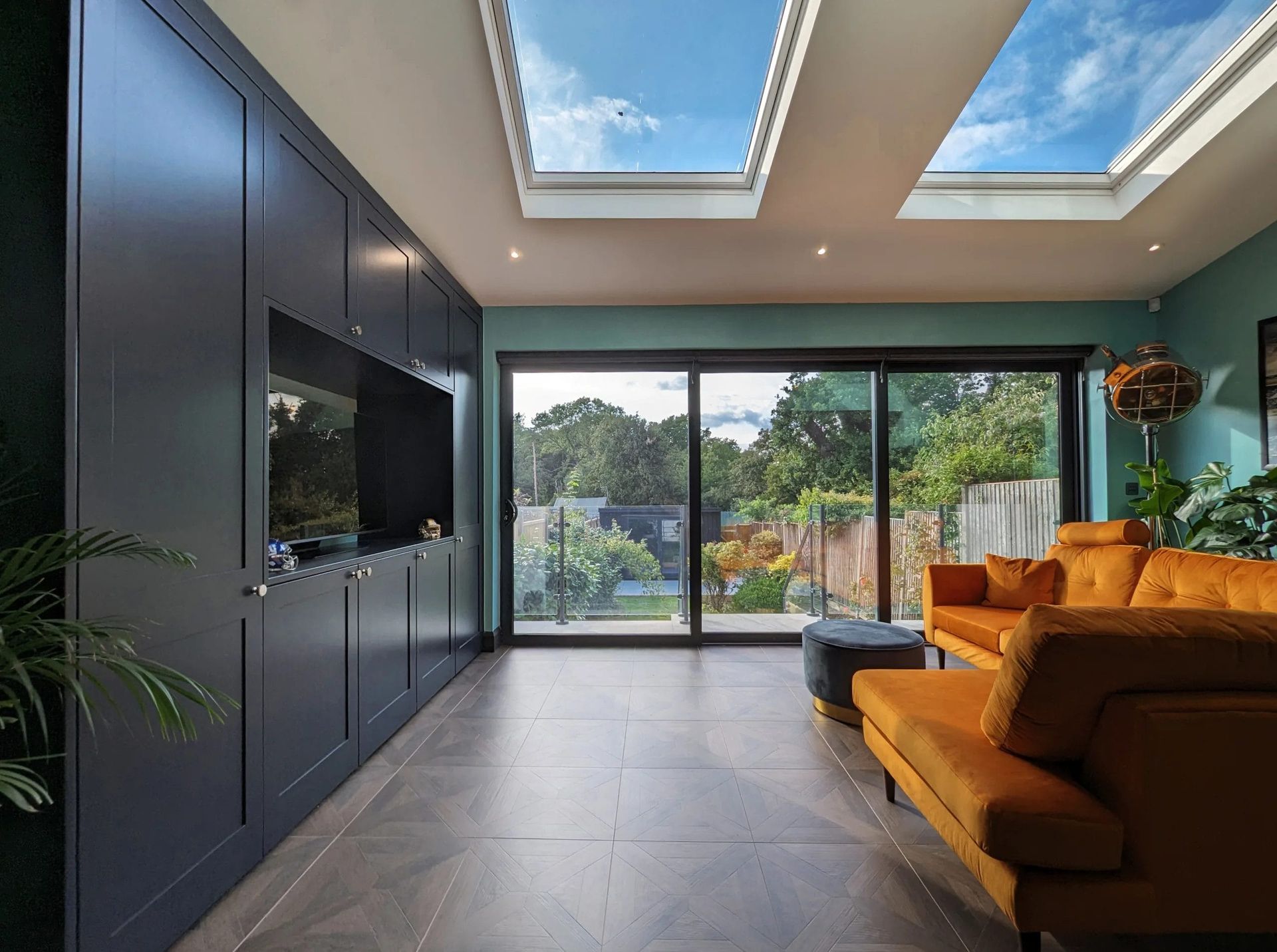 Living room with built-in dark blue cabinets, orange couch, large glass doors to a garden, and skylights showing a blue sky.