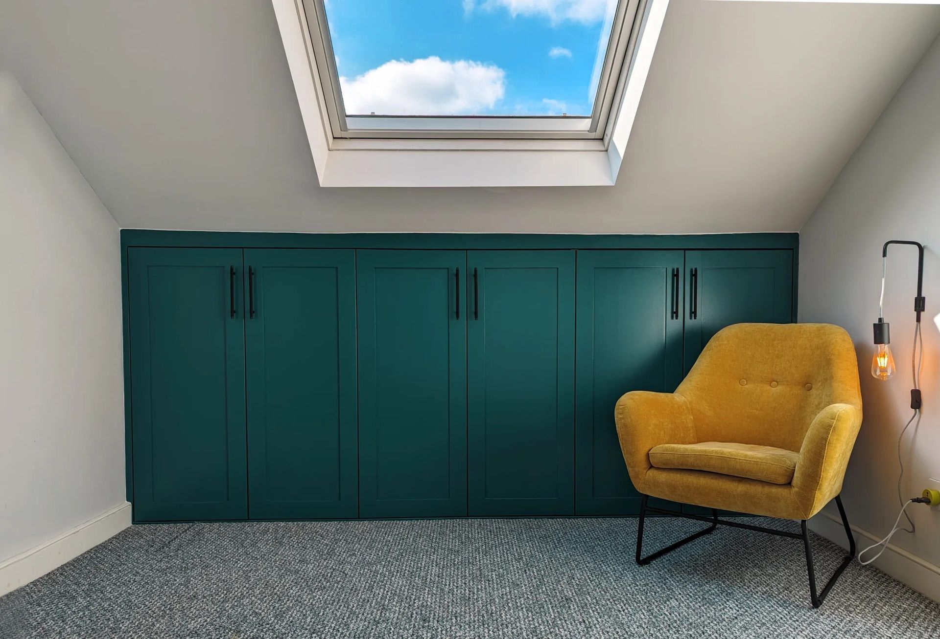 A room with green cabinets, yellow armchair, skylight, and gray carpet.