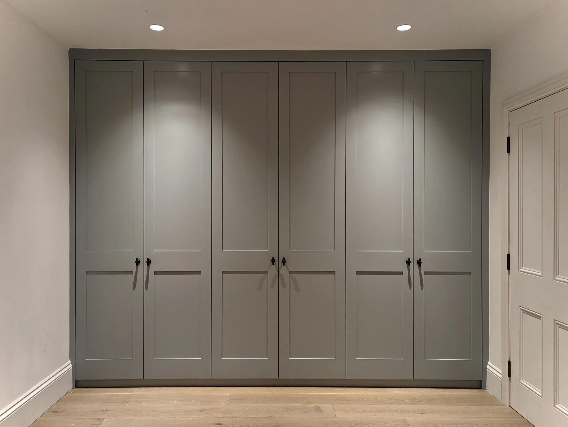 Gray built-in wardrobe with six doors, black handles, and recessed lighting in a room with a wood floor.