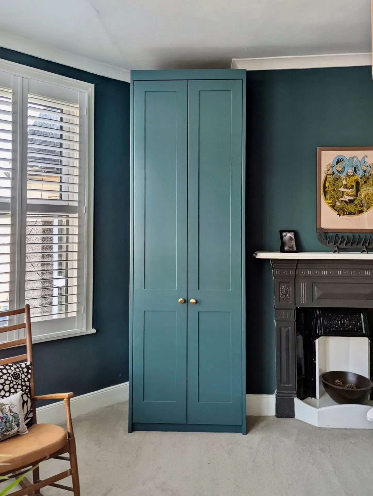Tall teal wardrobe between a window and fireplace in a teal-painted room with cream-colored flooring.