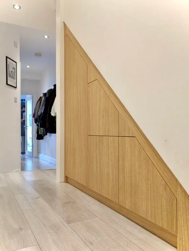 Built-in oak storage cabinets under stairs in a hallway with light wood flooring.