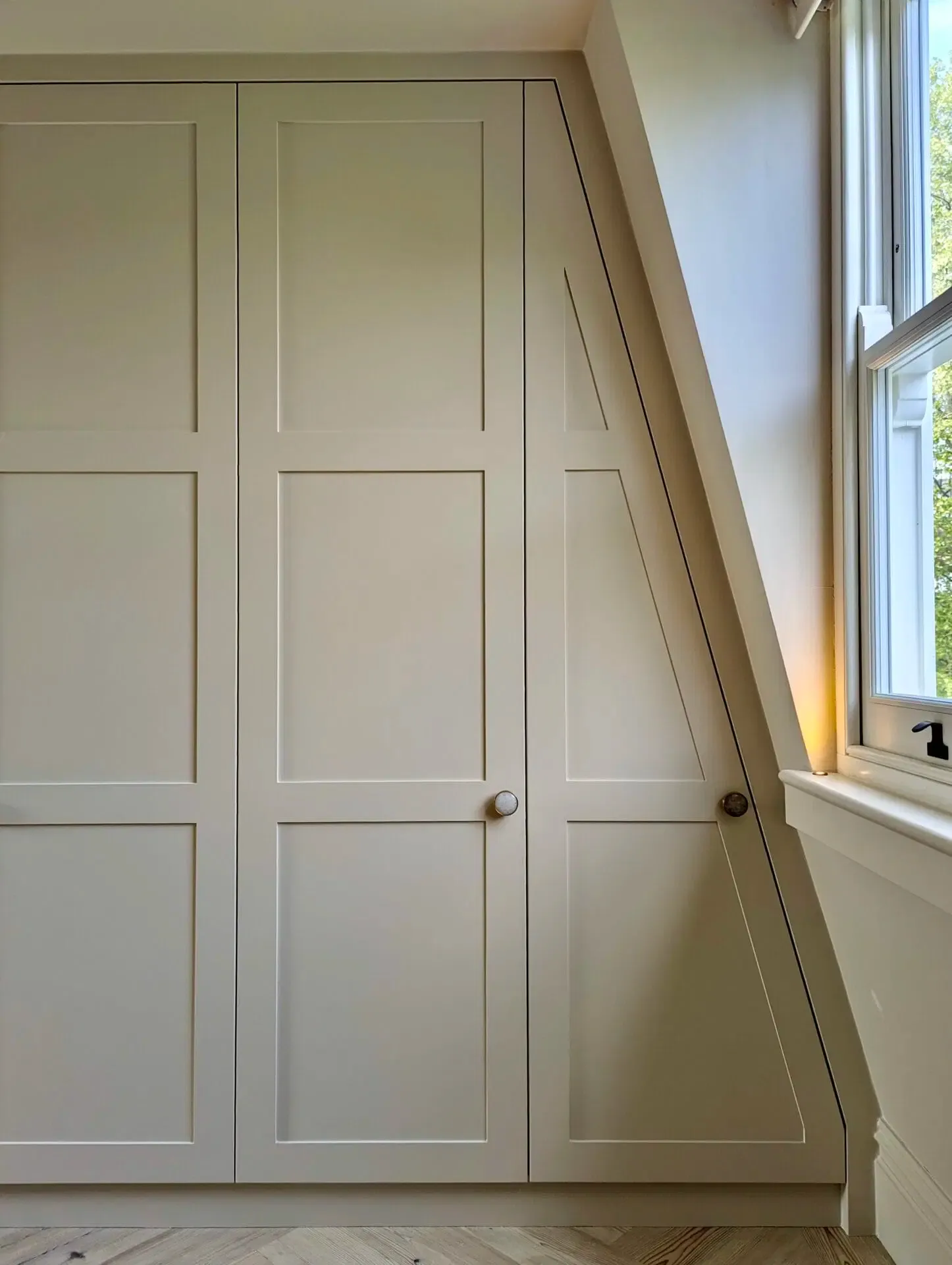 Built-in cream-colored wardrobe with paneled doors next to a window in a room.