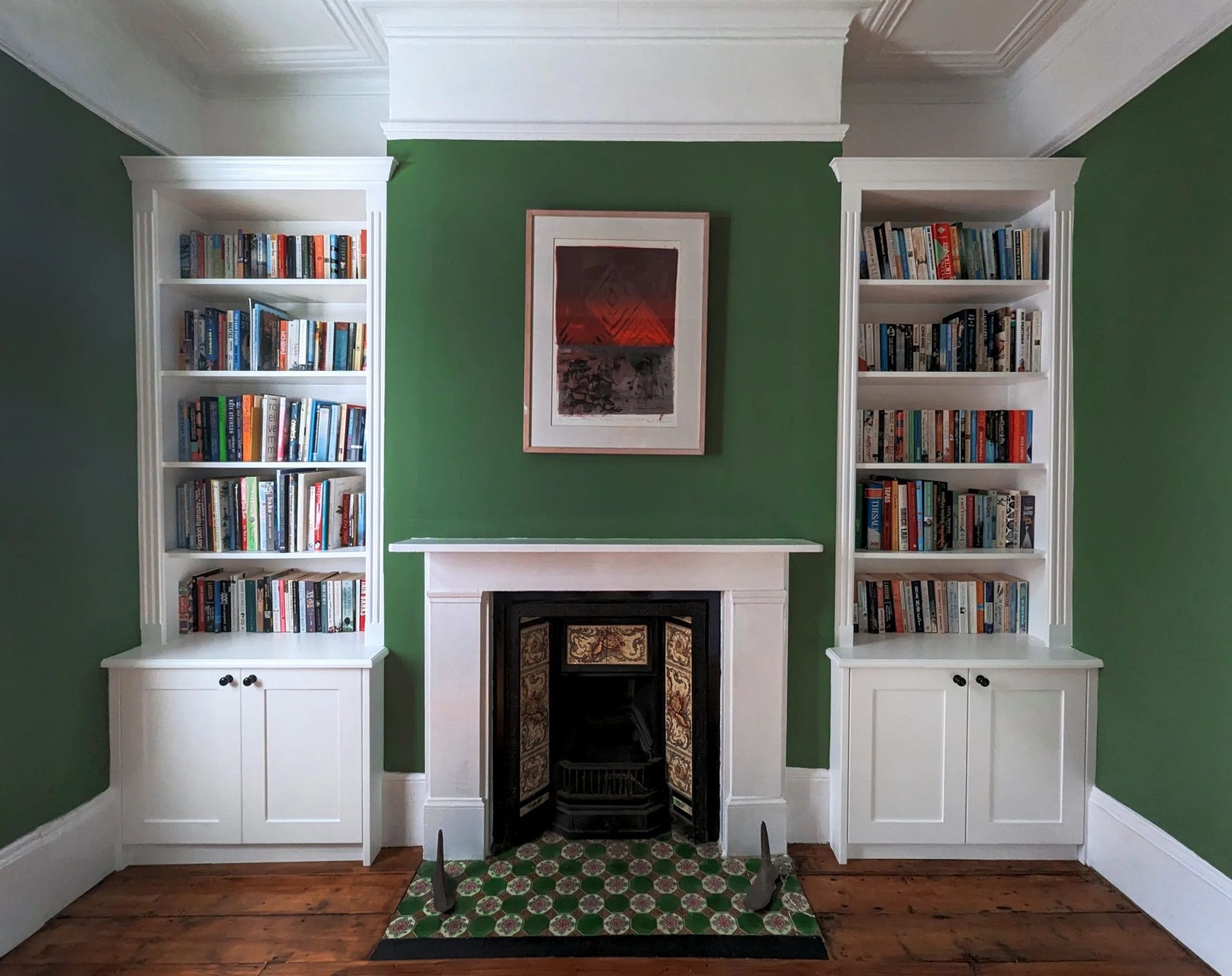 Green-walled room with white bookshelves flanking a fireplace with artwork above. Wooden floor.