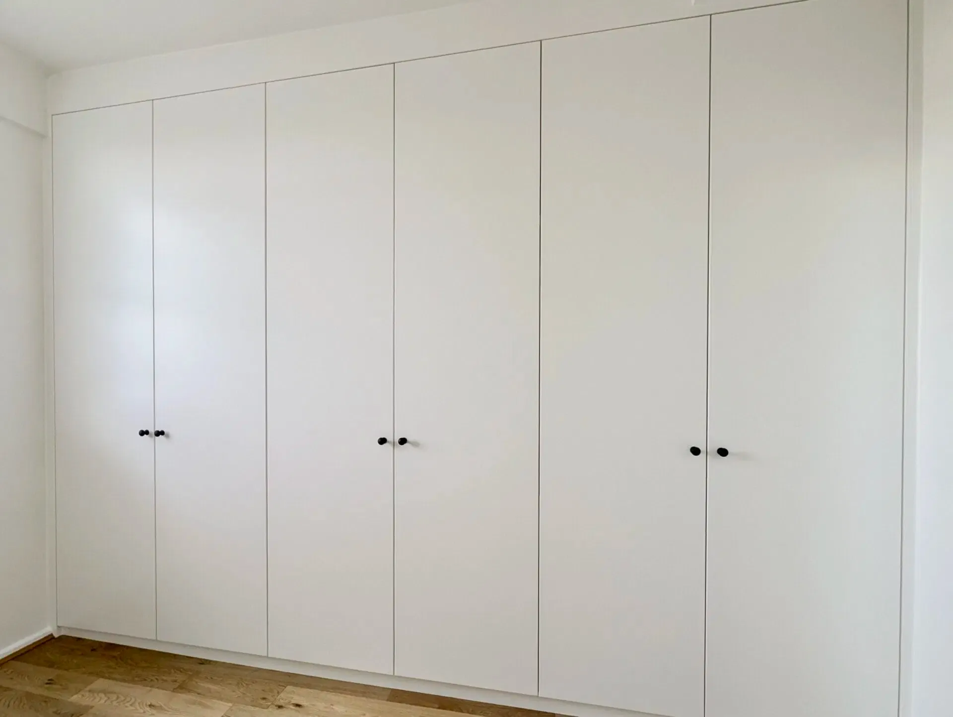 White built-in wardrobe with six tall, flat doors and small black handles, in a room with light wooden flooring.