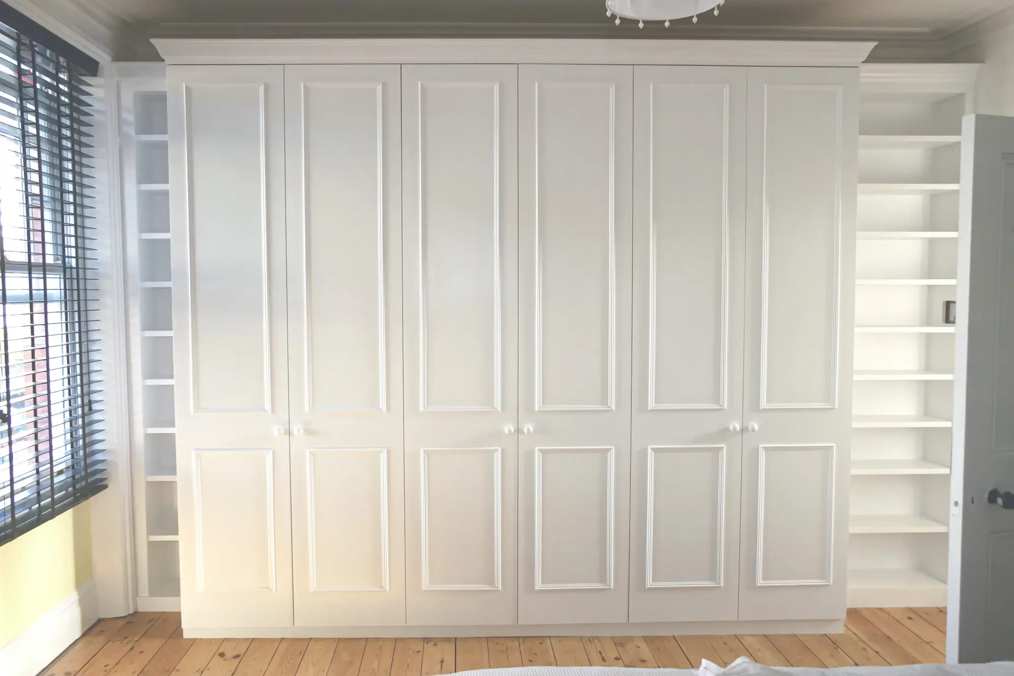 White built-in wardrobe with six doors, flanked by open shelving units in a room with a window.