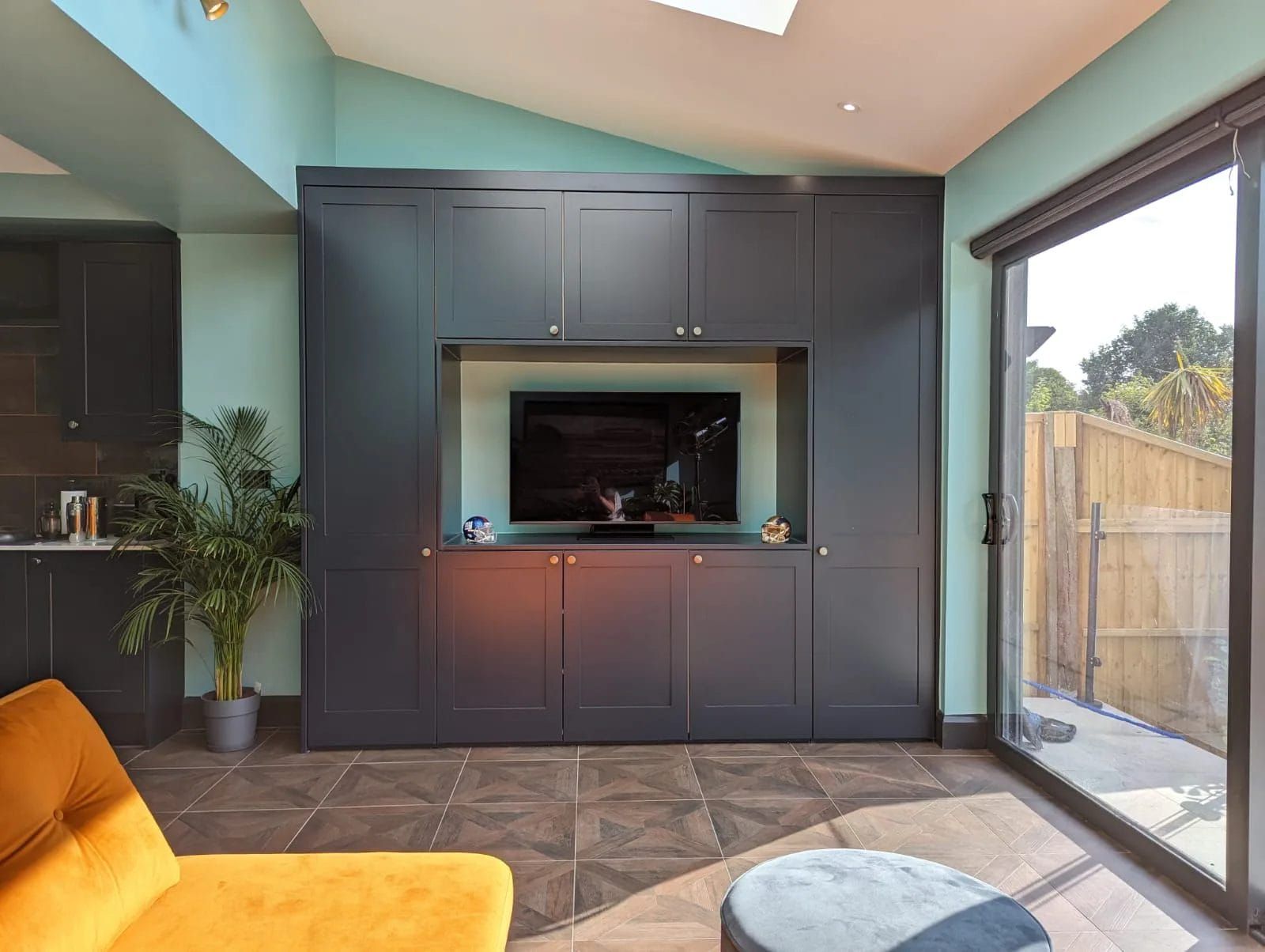 Built-in dark gray cabinet with TV in recessed area, teal wall, sliding glass door, yellow couch, and a plant.