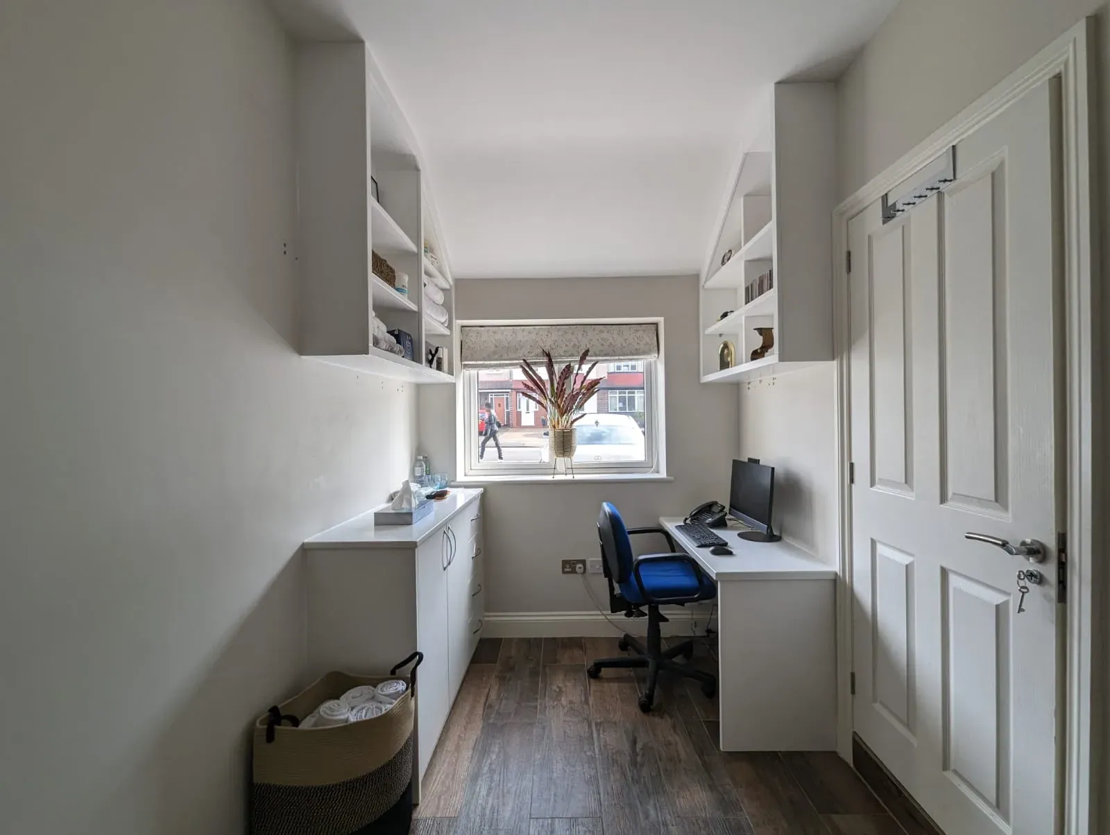 Home office with built-in desk, shelving, window, and door. Gray walls, white cabinets, and wood-look flooring.