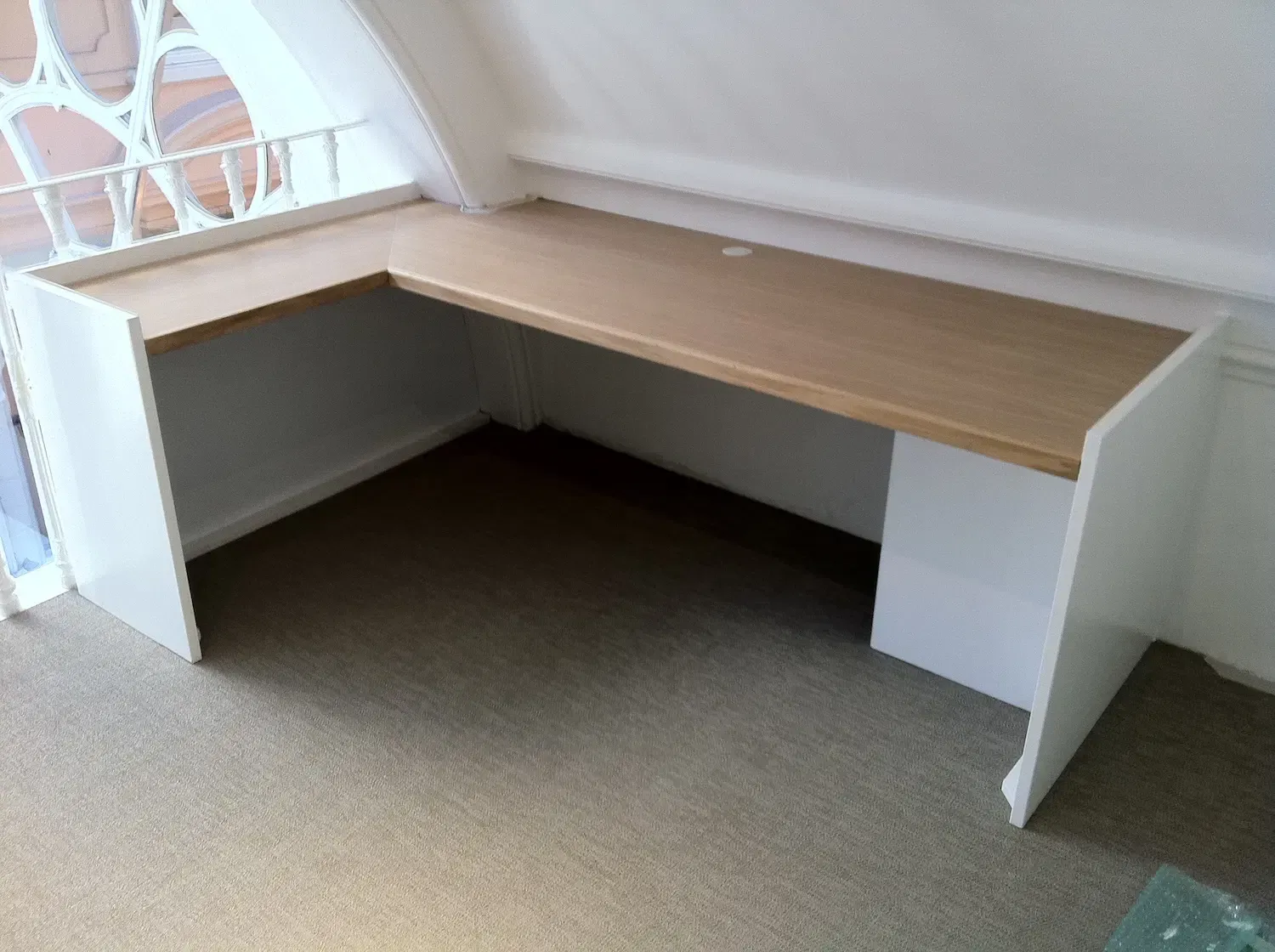 Built-in L-shaped desk with a light wood top and white sides, in a room with a sloping ceiling and a window.