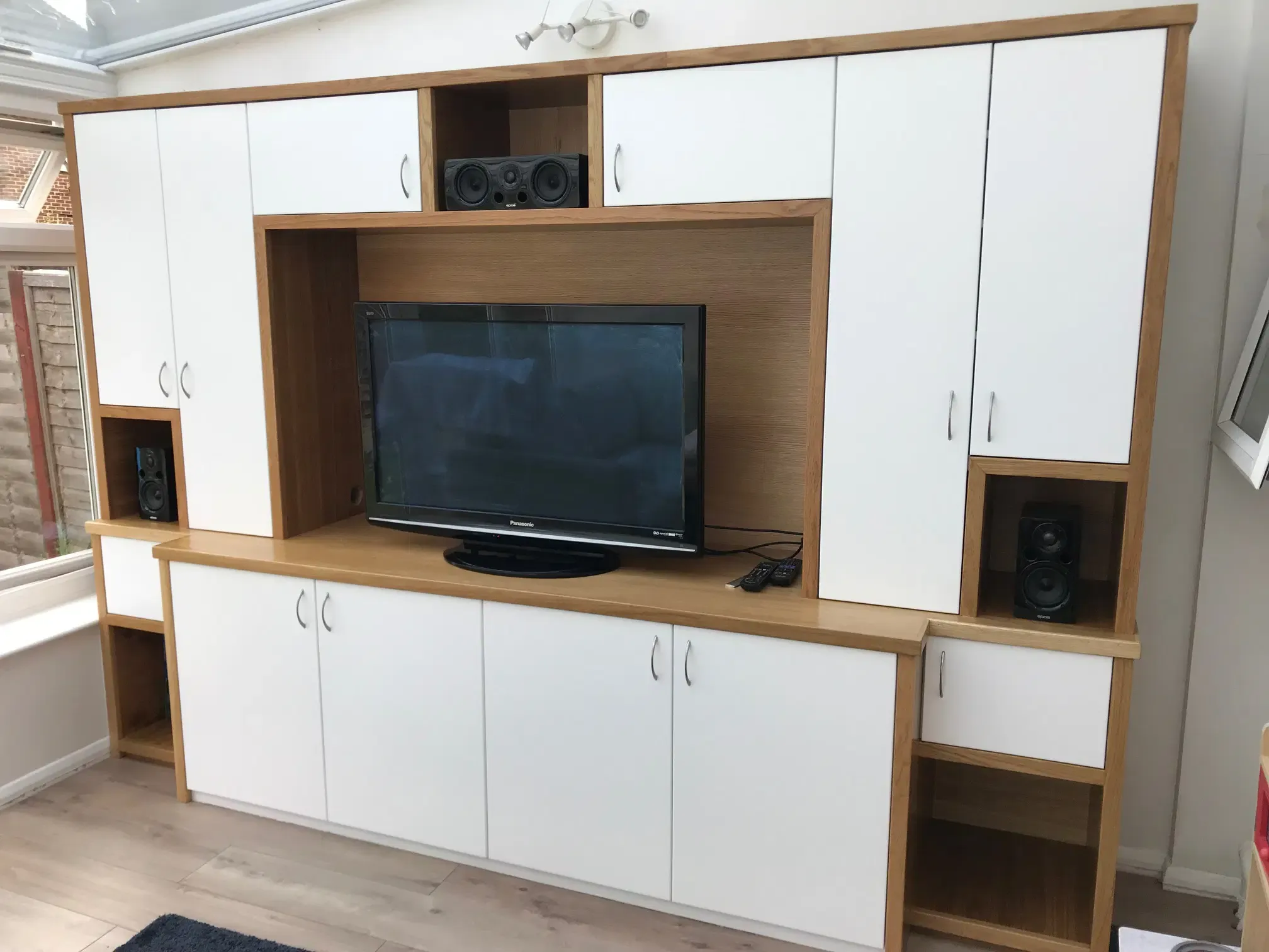 Built-in white cabinets with wooden accents surround a flat-screen TV and speakers in a well-lit room.
