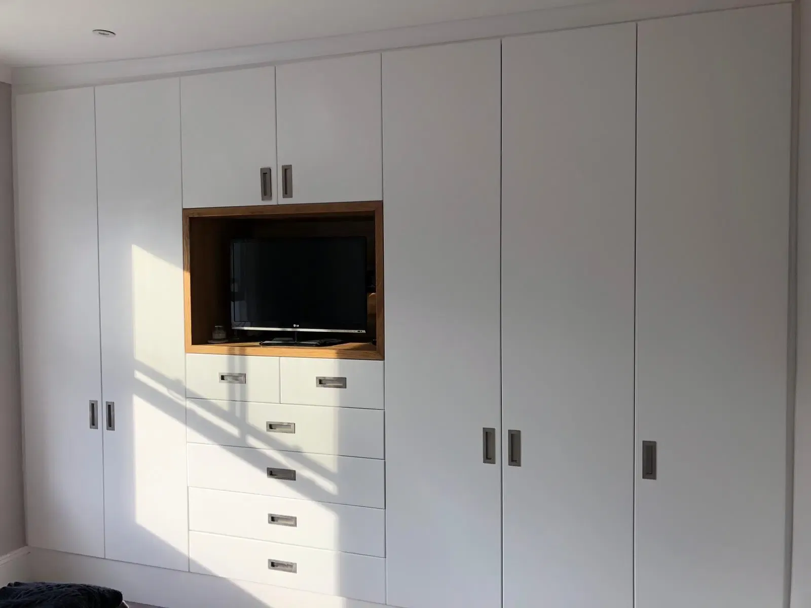 White built-in closet with centered TV, drawers, and wood-toned frame, with dark handles and a light wall.