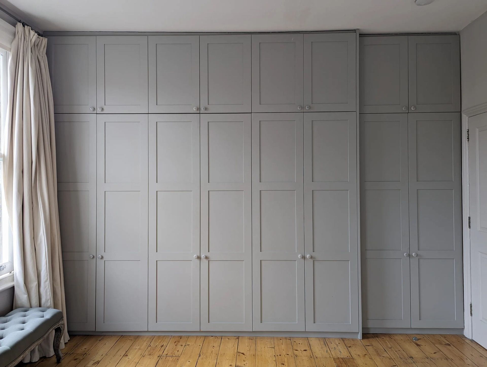 Gray built-in wardrobes cover a wall. Beige curtains frame a window. Light wood floor.