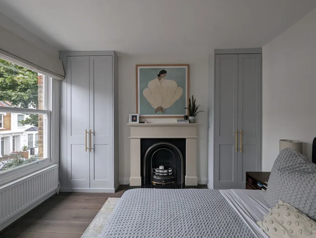 Bedroom with fireplace, built-in closets, and window with a view. Light blue cabinets, hardwood floors, and a gray bedspread.