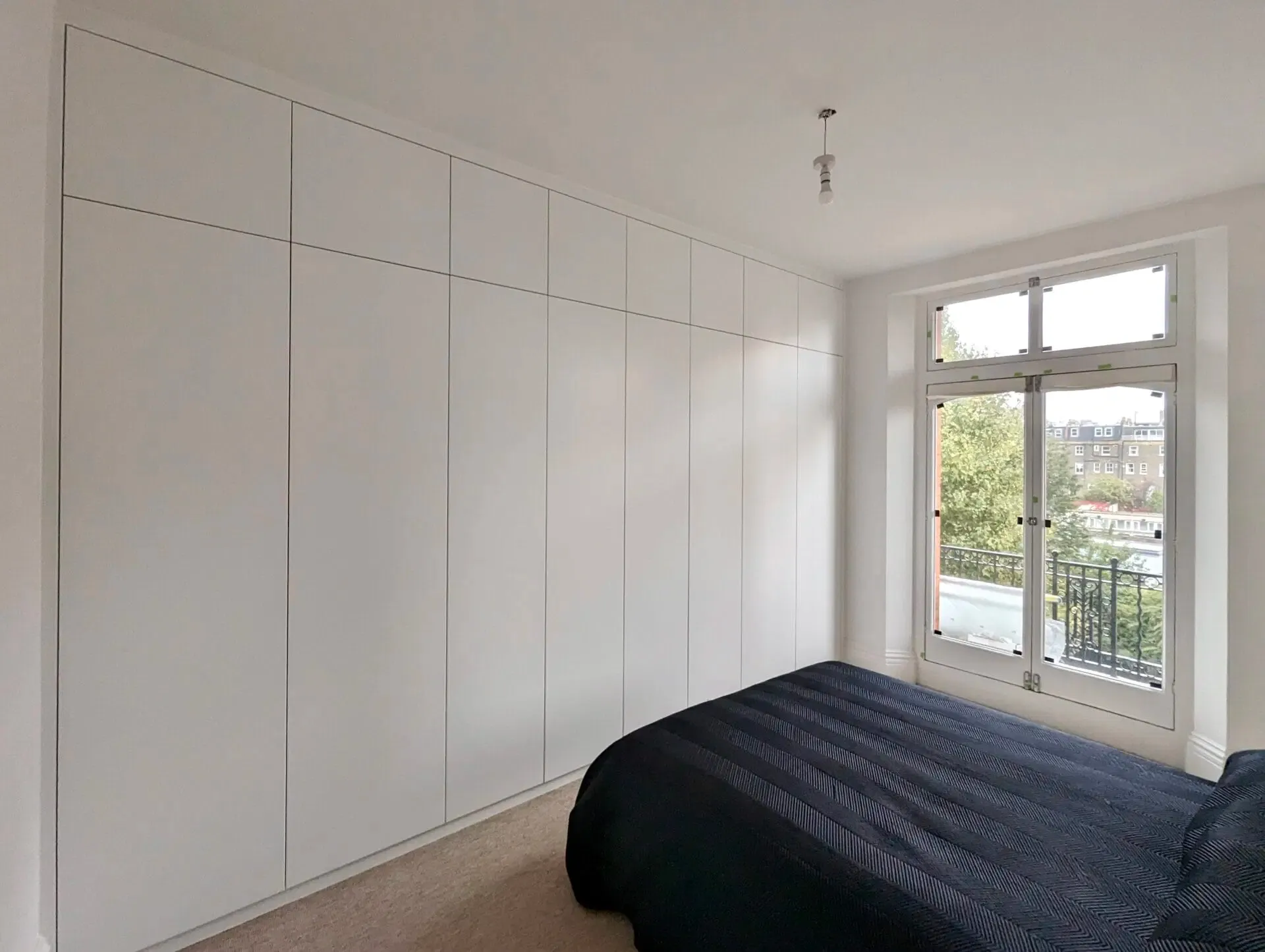 White built-in wardrobe dominates a bedroom with a bed and window overlooking greenery.