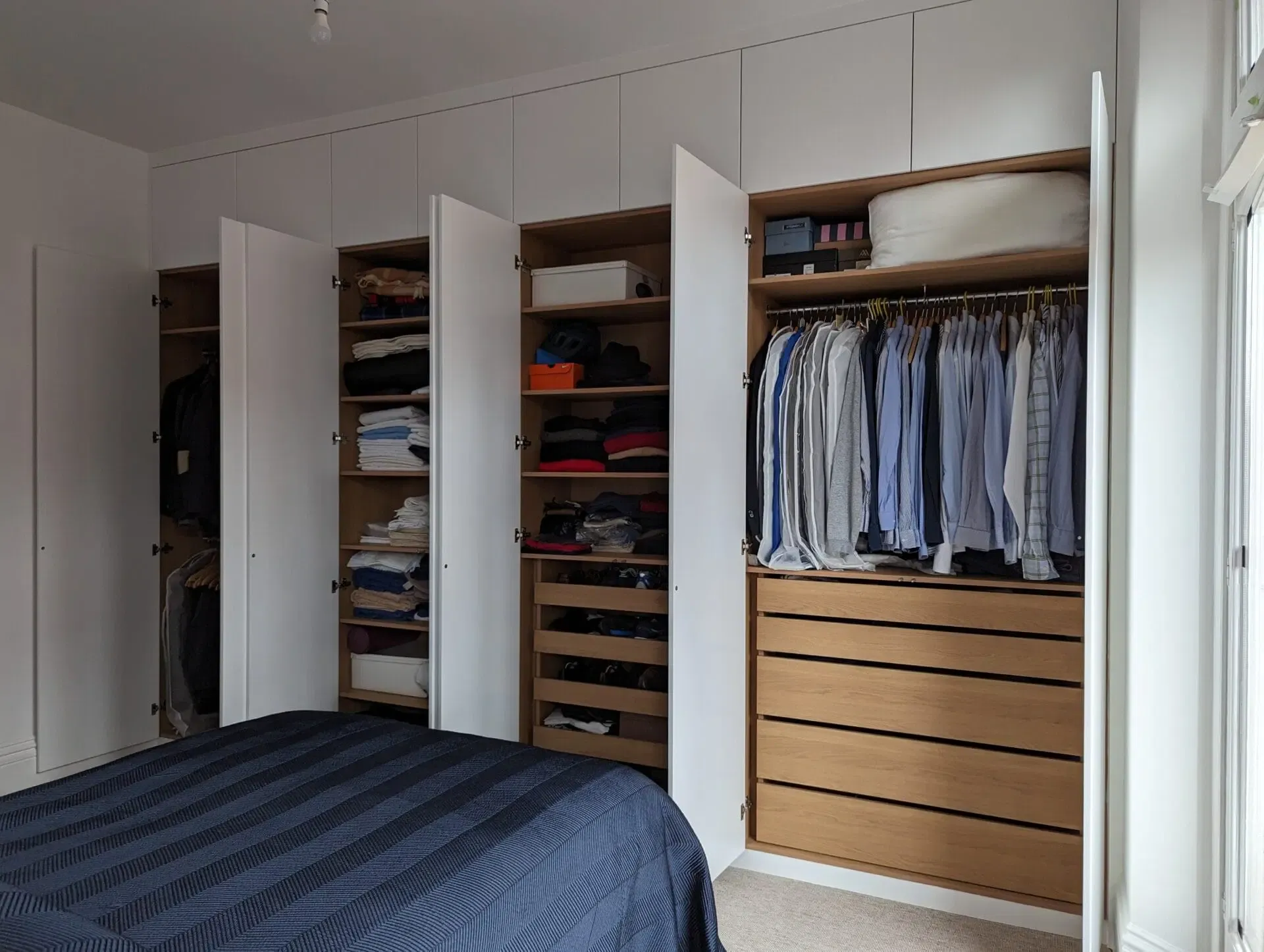 Bedroom closet with white doors open to reveal clothes and drawers; bed in foreground.