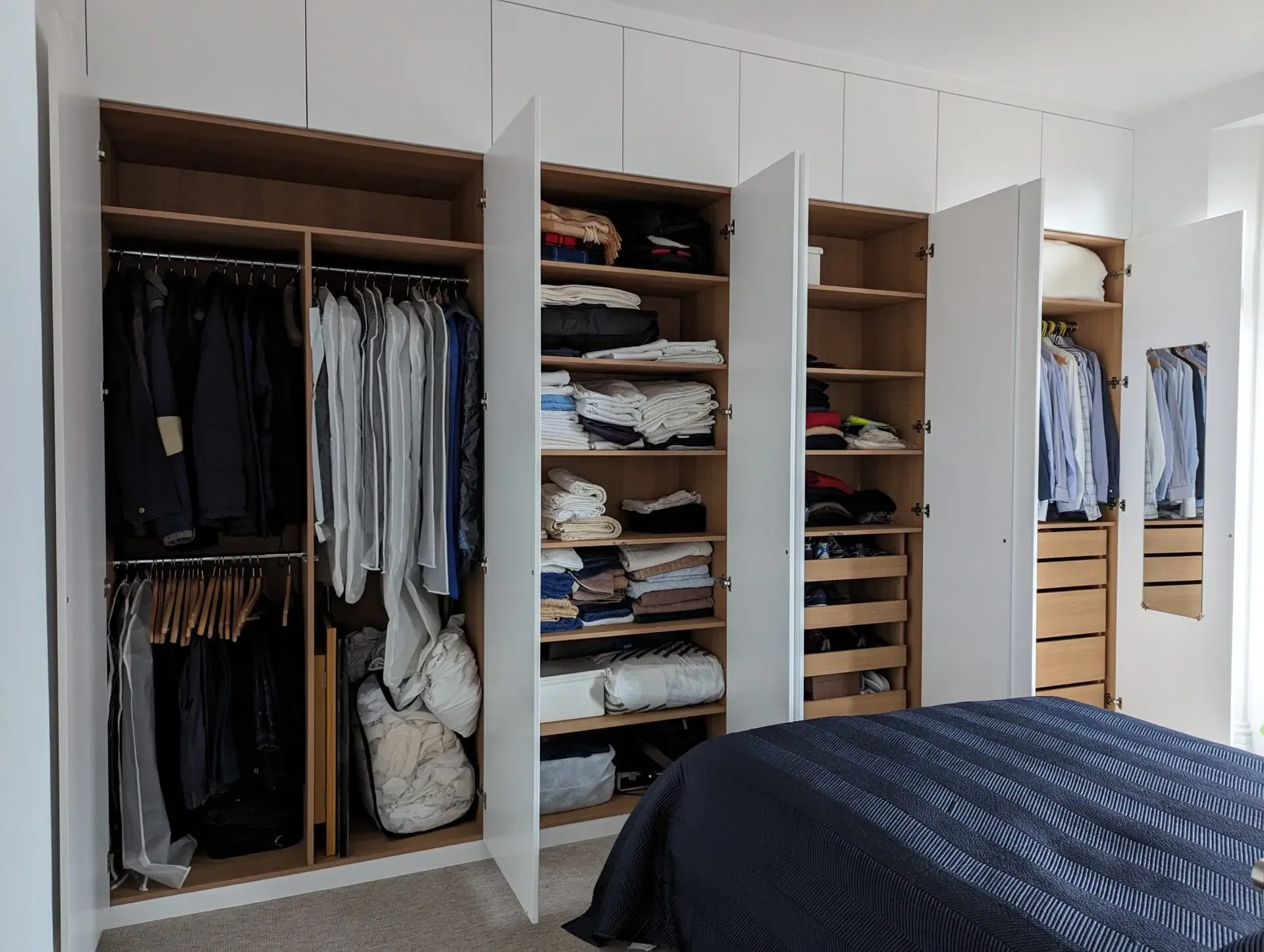 Wooden closet with open doors, revealing clothes and shelves. White doors, neutral-toned interior, bed in foreground.