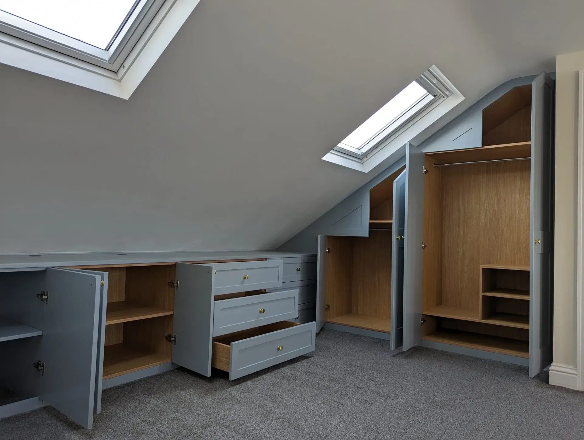 Gray built-in cabinets under sloped ceiling with skylights, featuring open doors and drawers; carpeted floor.