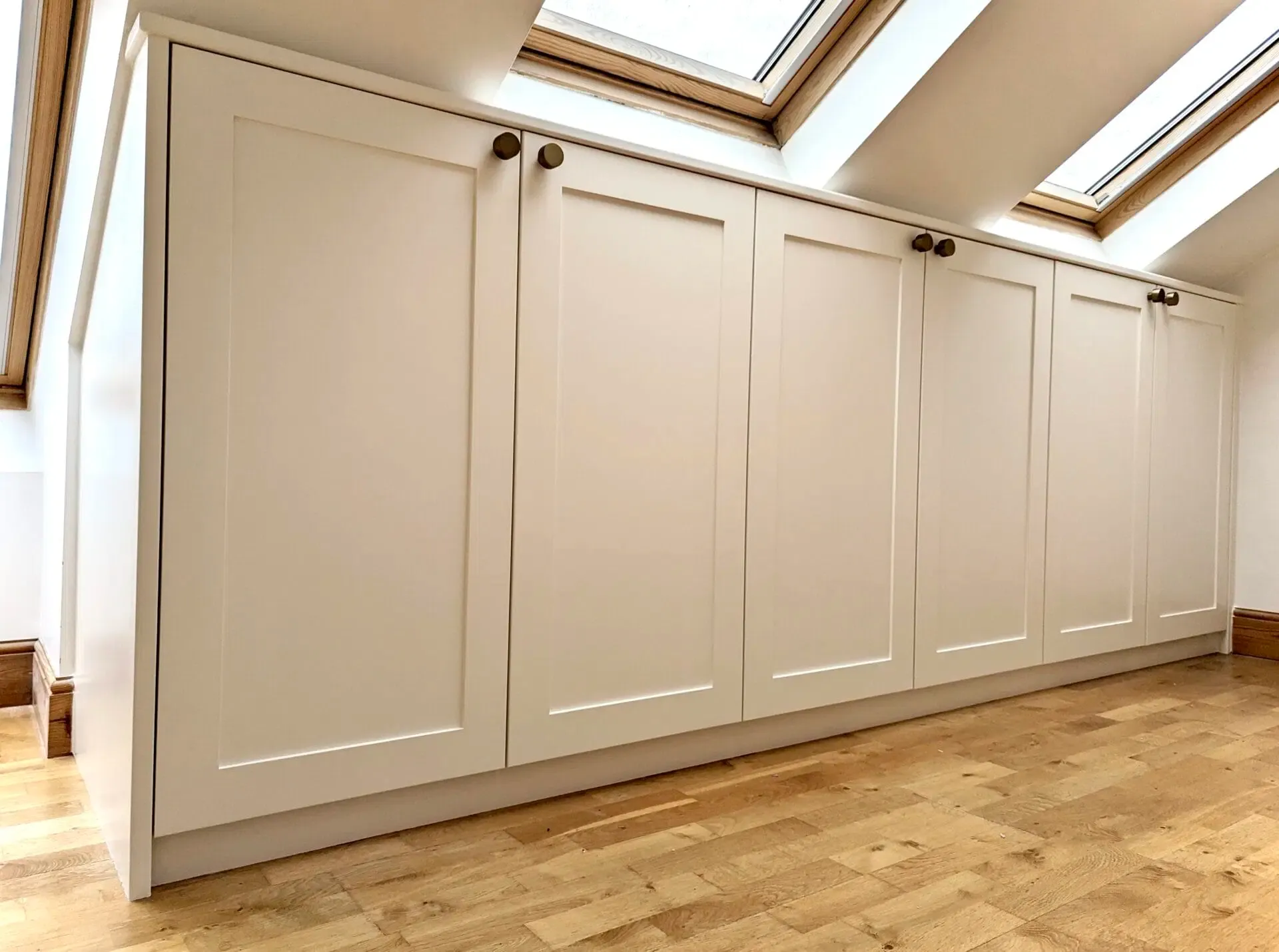 Built-in cream-colored cabinets with dark handles, along an angled wall with skylights, above wood flooring.