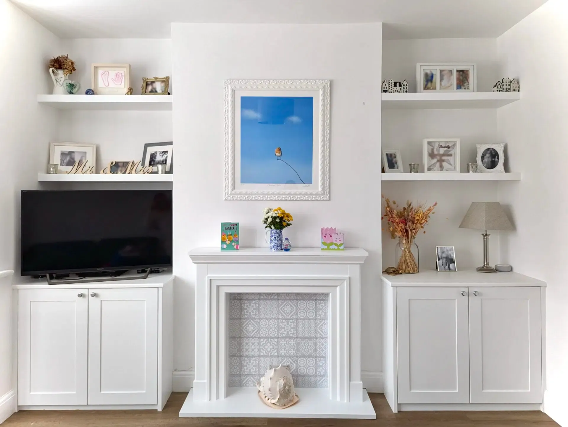 Built-in white shelves and cabinets flank a fireplace, with a framed artwork and TV, all in a bright, modern interior.