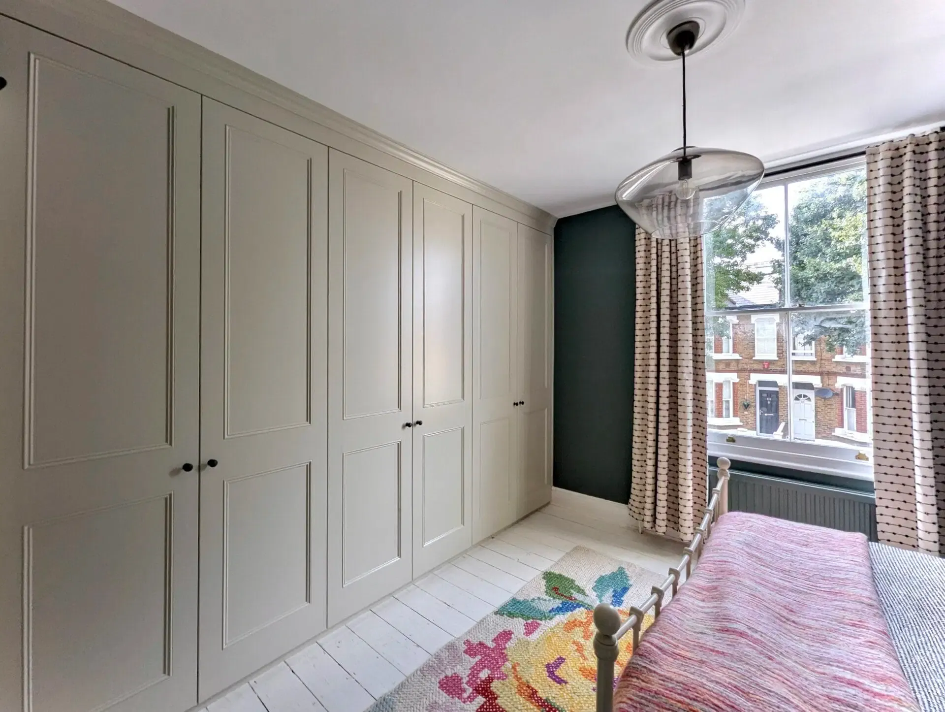 Bedroom with built-in gray wardrobes, floral rug, window with curtains, and hanging light fixture.