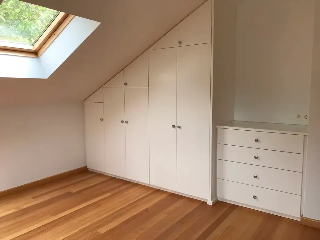 A bright room with built-in white cabinets under a sloped ceiling and wooden floors.