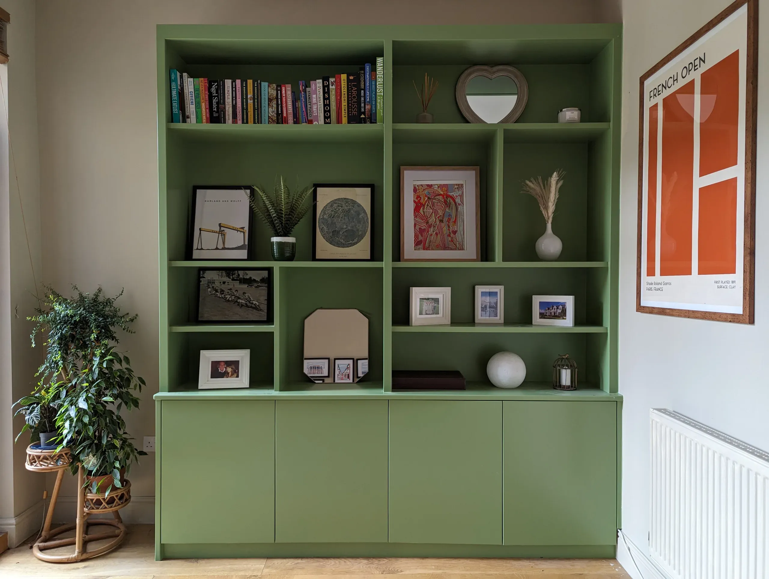 Green built-in bookcase with books, framed art, and decorative objects. A plant sits nearby, beside a light-colored wall.