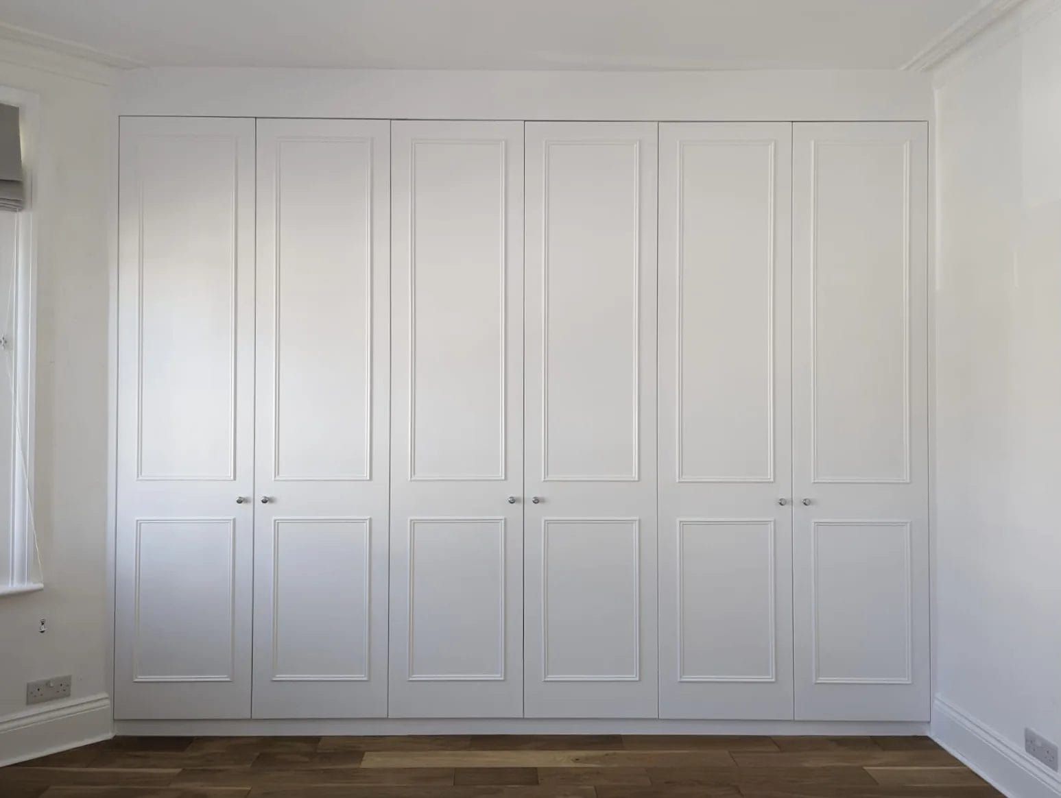 White built-in wardrobe with six doors in a room with a hardwood floor and window.