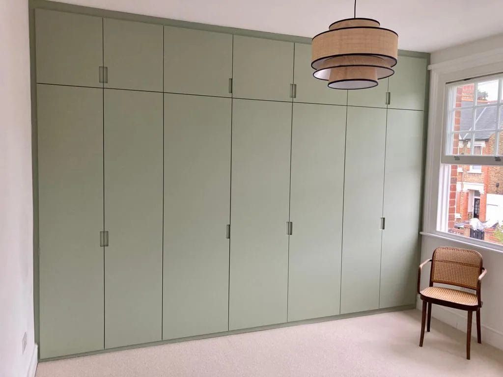 Green built-in closet in bedroom with woven chair and window, beige carpet, and decorative pendant light.
