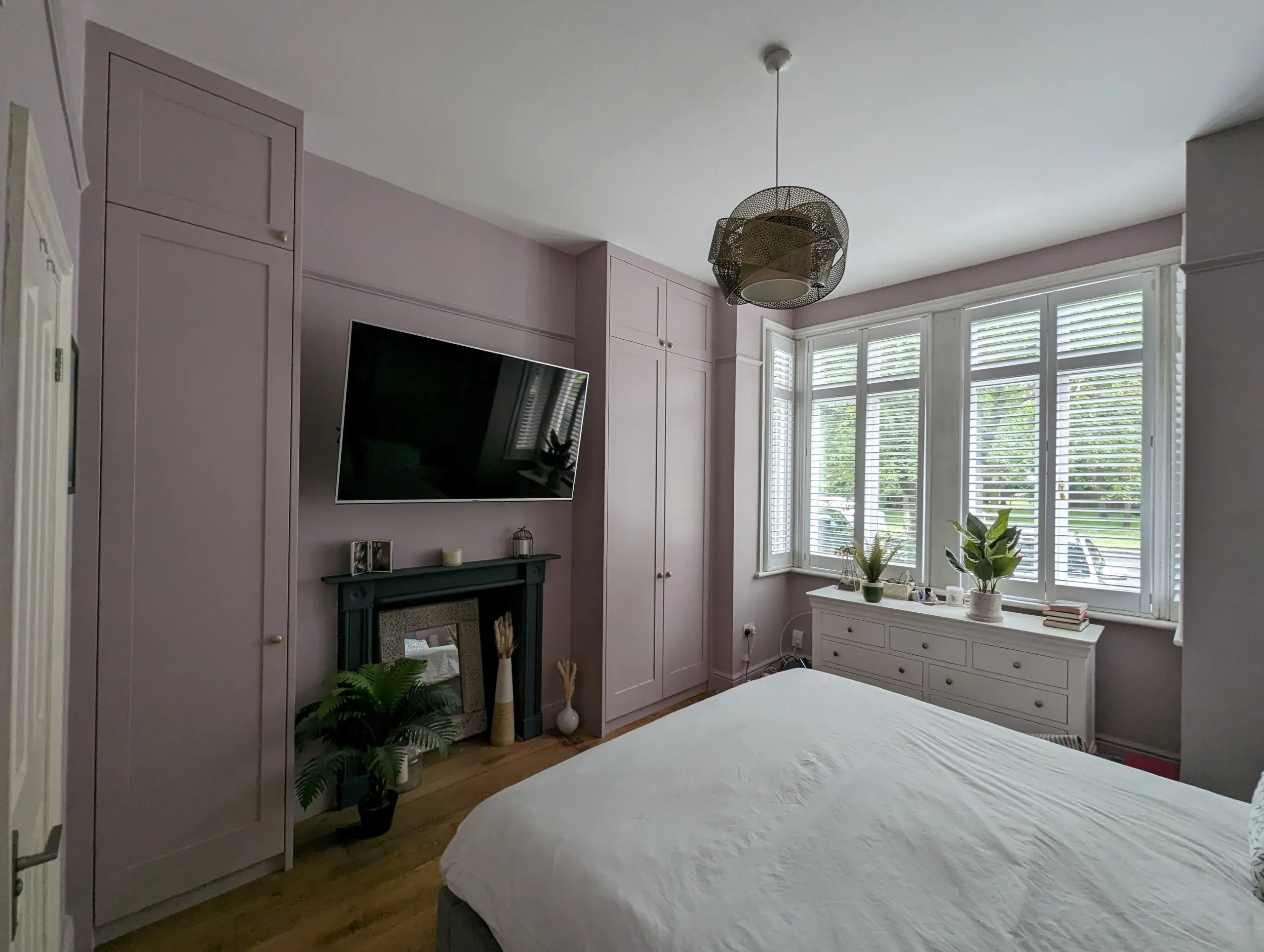Bedroom with lilac walls, built-in wardrobes, fireplace, TV, white dresser, large window with shutters, and bed.