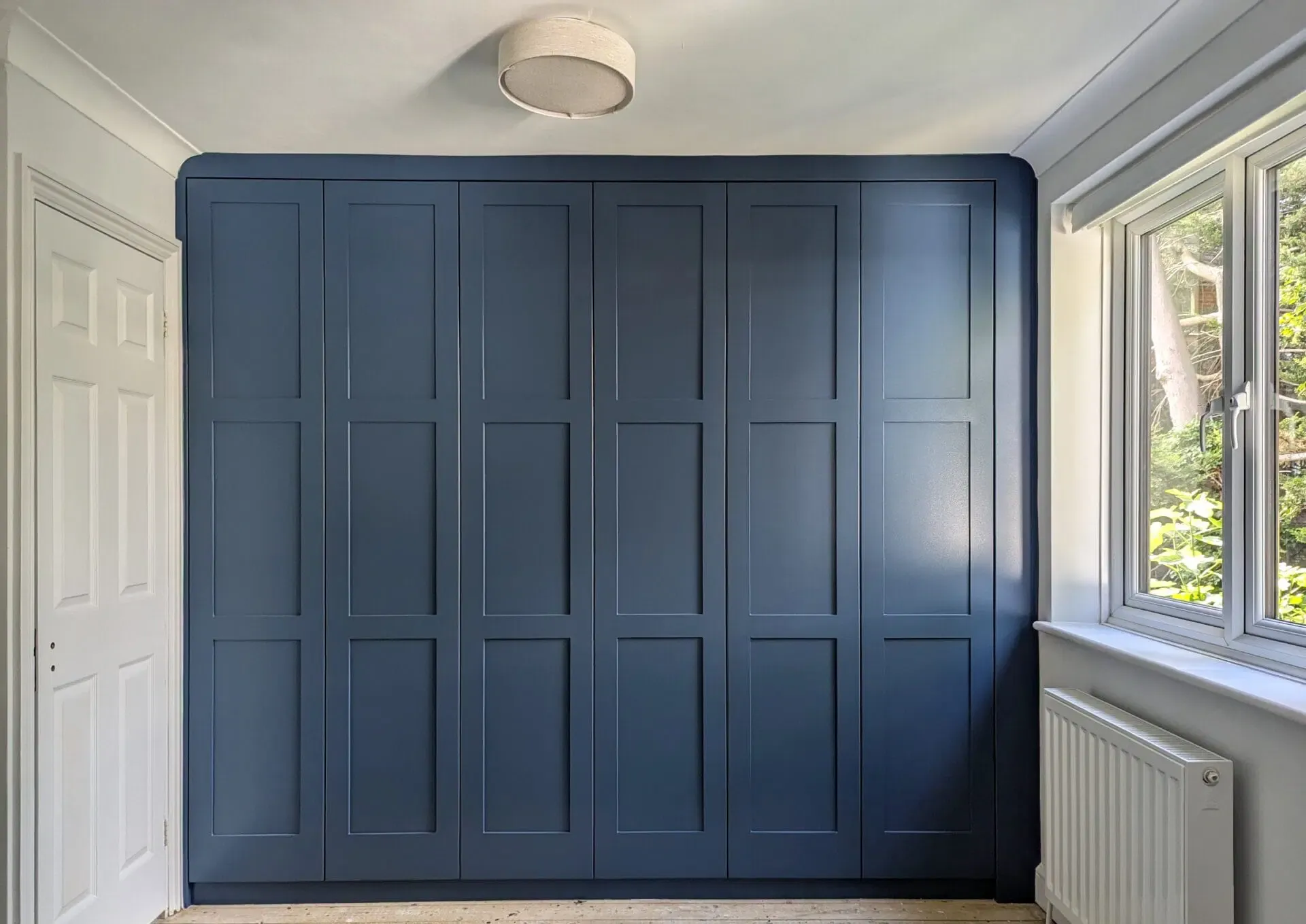 Blue built-in wardrobe with six doors, next to a window with a radiator below and a white door on the left.