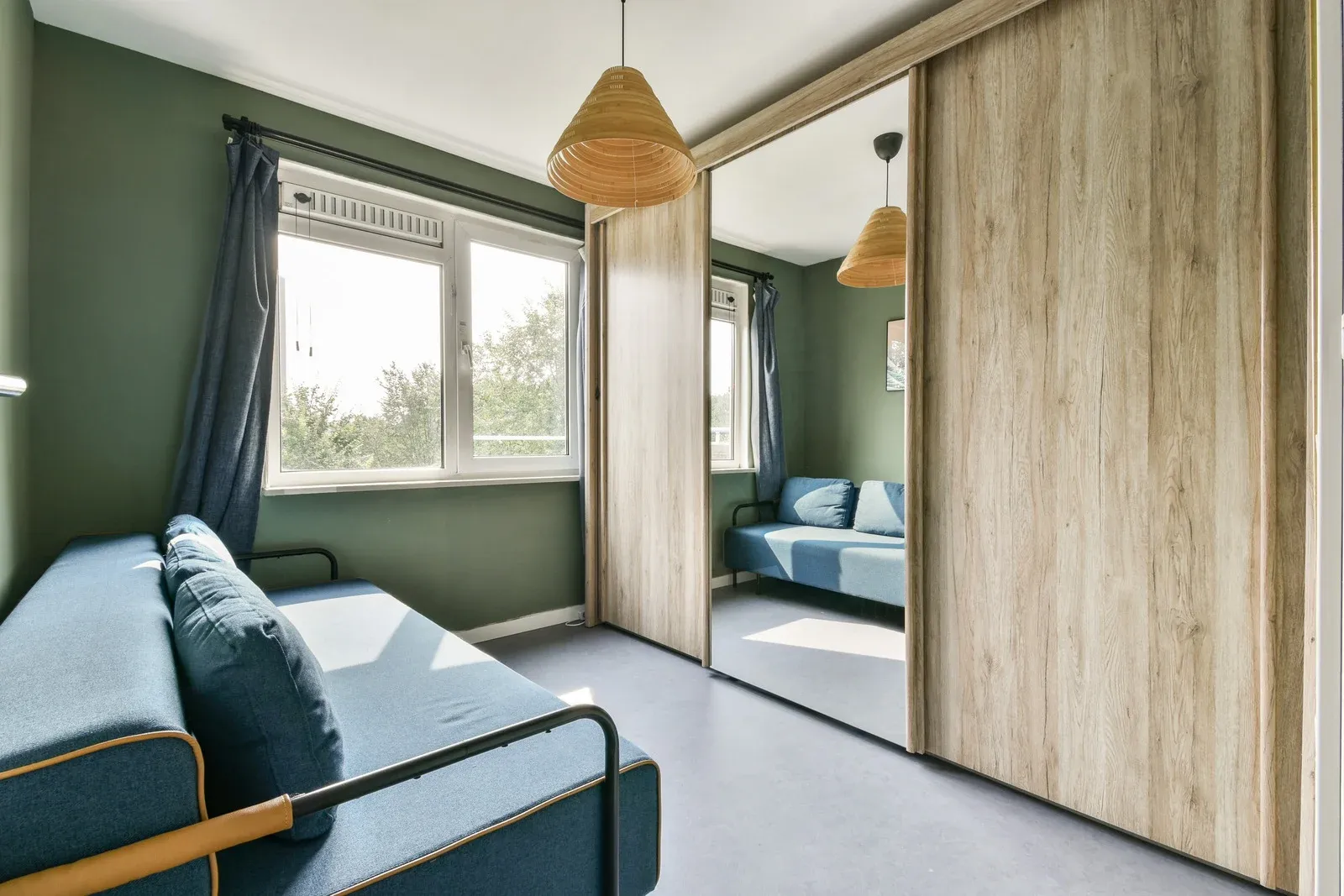 Room with blue sofa, green wall, window with curtains, and mirrored closet. Light fixture.