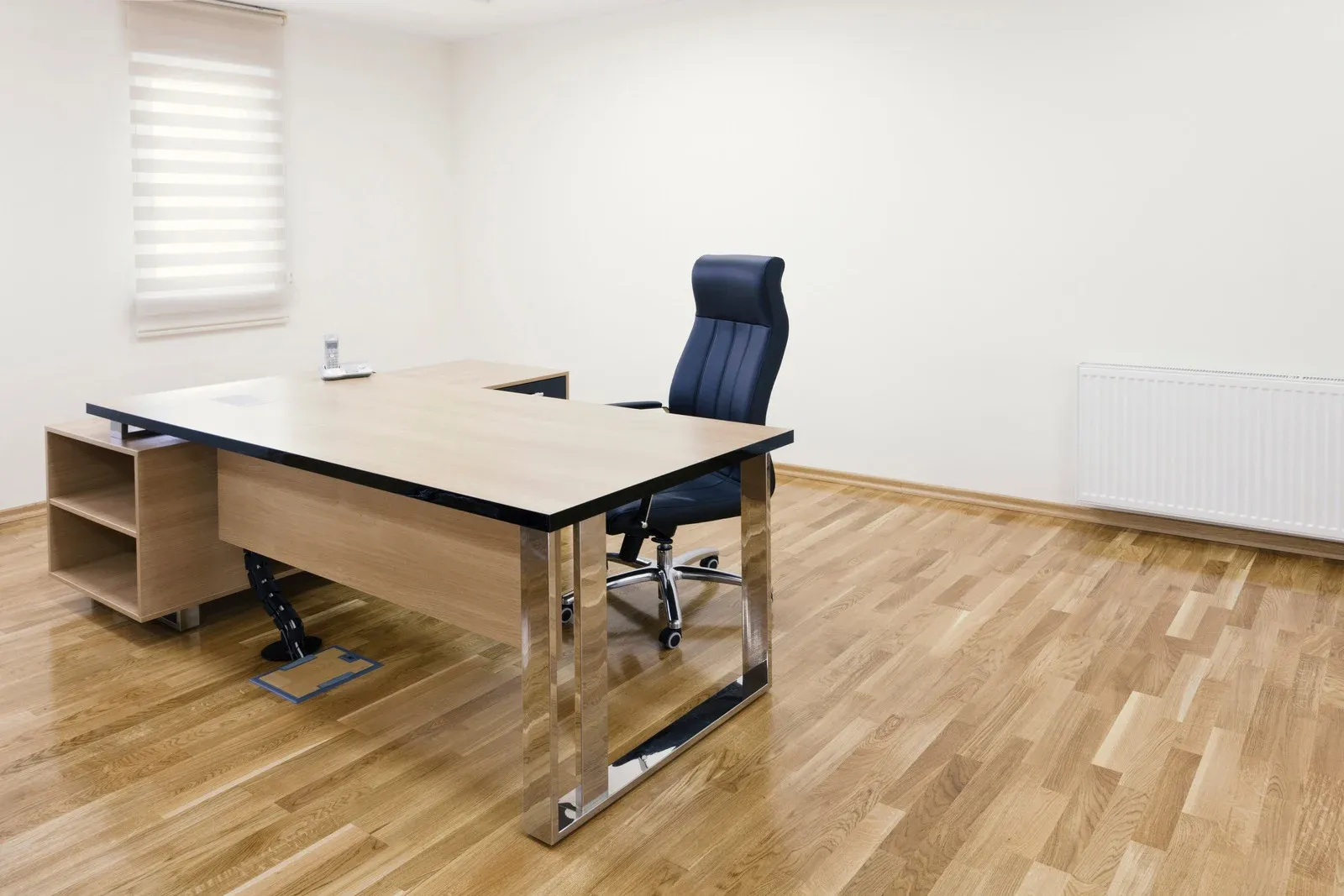 Empty office with wooden floor, desk, and chair. Window with blinds, radiator on the wall.