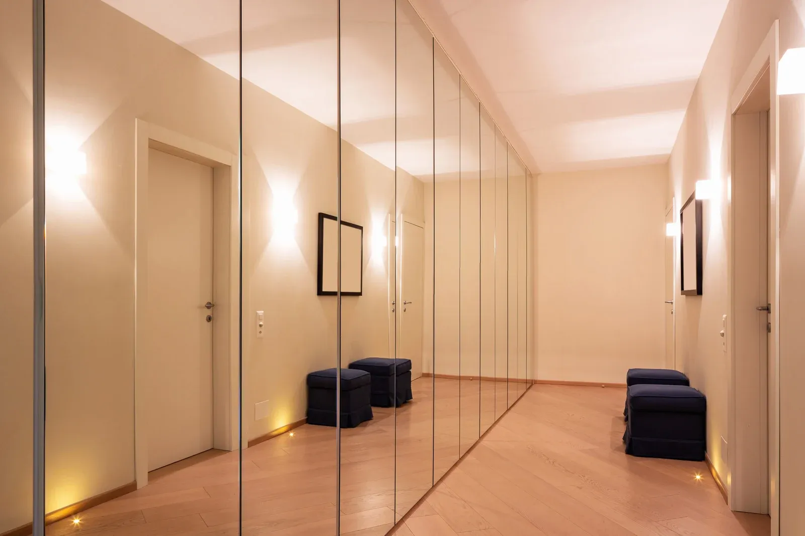 Hallway with full-length mirrored closet doors, a wooden floor, and two dark blue ottomans.