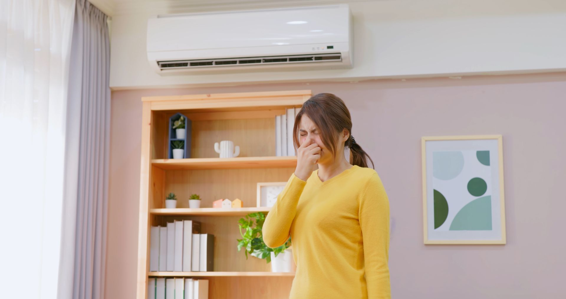A woman is covering her mouth while standing in front of an air conditioner in a living room.