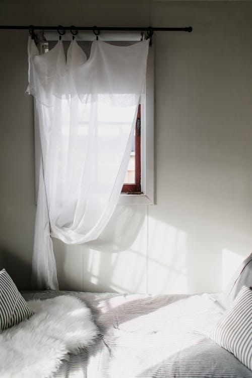 Sunlit bedroom corner with sheer white curtains, a small window, and a bed with striped pillows.