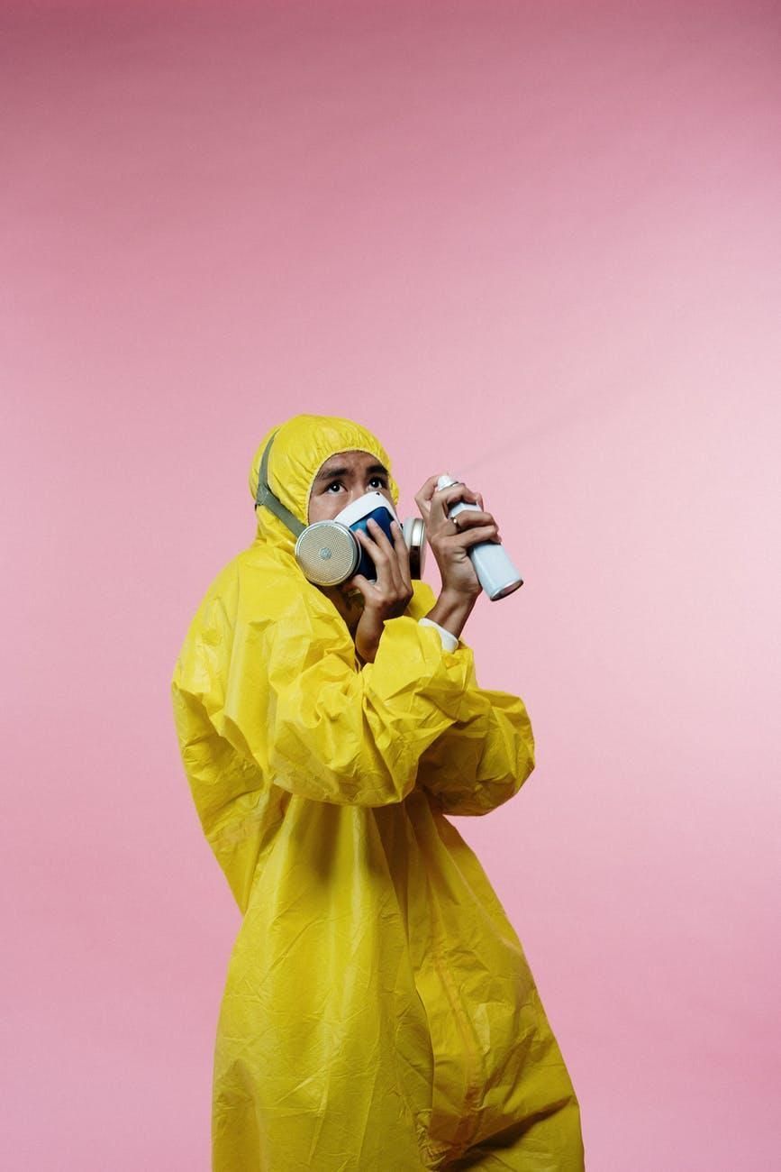 Person in yellow hazmat suit wearing a respirator and holding a spray can against a pink background