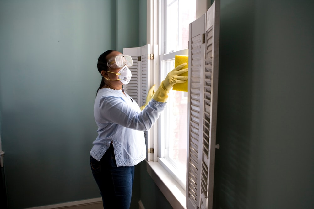 Person in gloves and mask cleaning a window with a spray bottle indoors