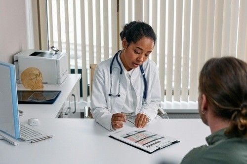 A medical professional in a white coat with a stethoscope discusses a document with a patient in an office.