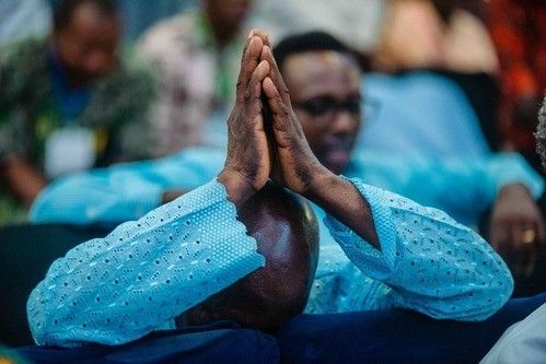 Man in light blue shirt with hands clasped in prayer, blurred background.