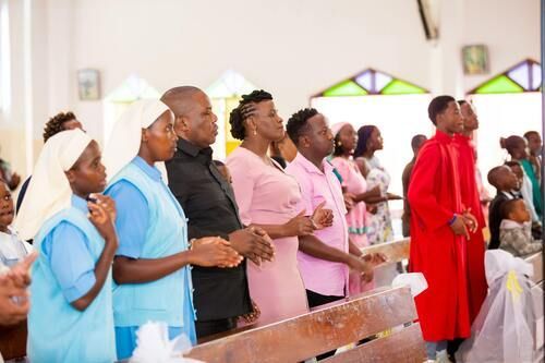 People standing in a church, some wearing light blue uniforms, others in formal attire, some clapping.