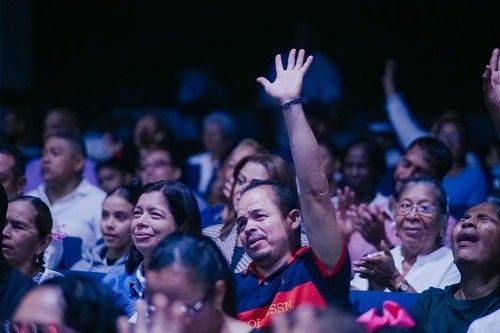 People in a brightly lit auditorium, some with hands raised in praise.