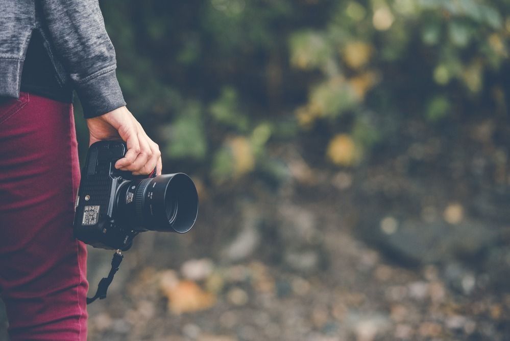 A person in a gray sweater and maroon pants holds a black DSLR camera against a blurred outdoor background.